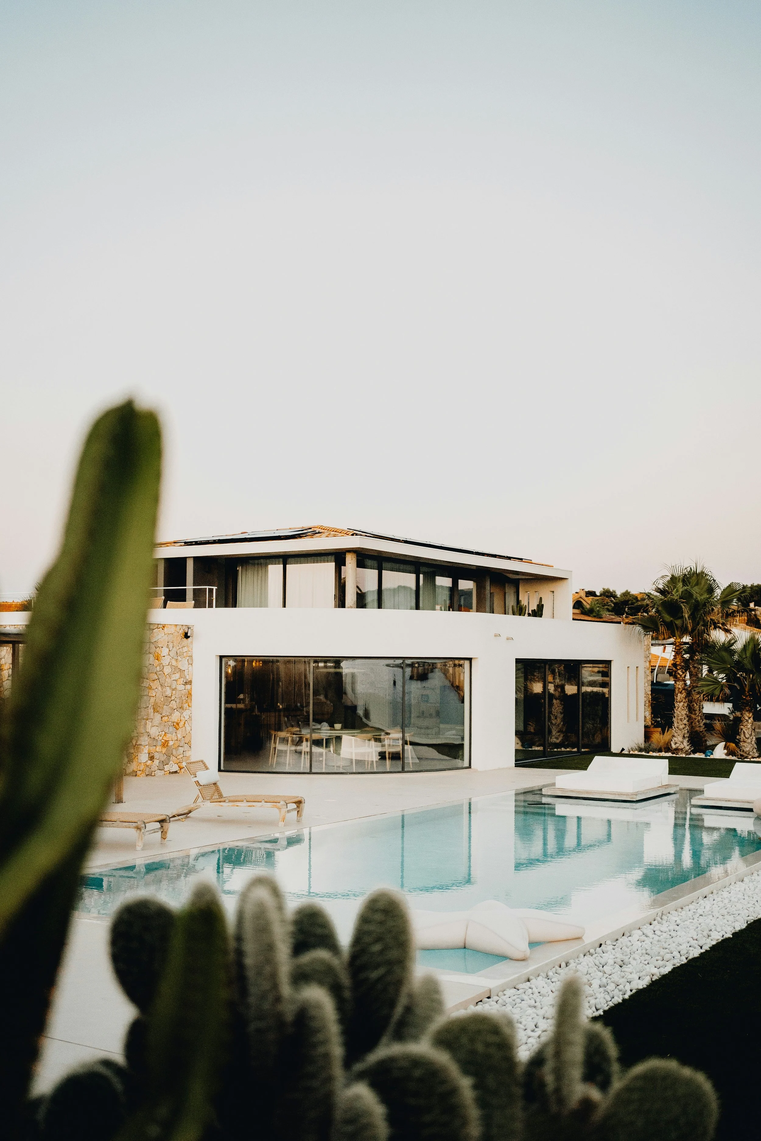 Modern white house with large glass windows and a pool in the backyard, surrounded by desert plants and palm trees, under a clear sky.