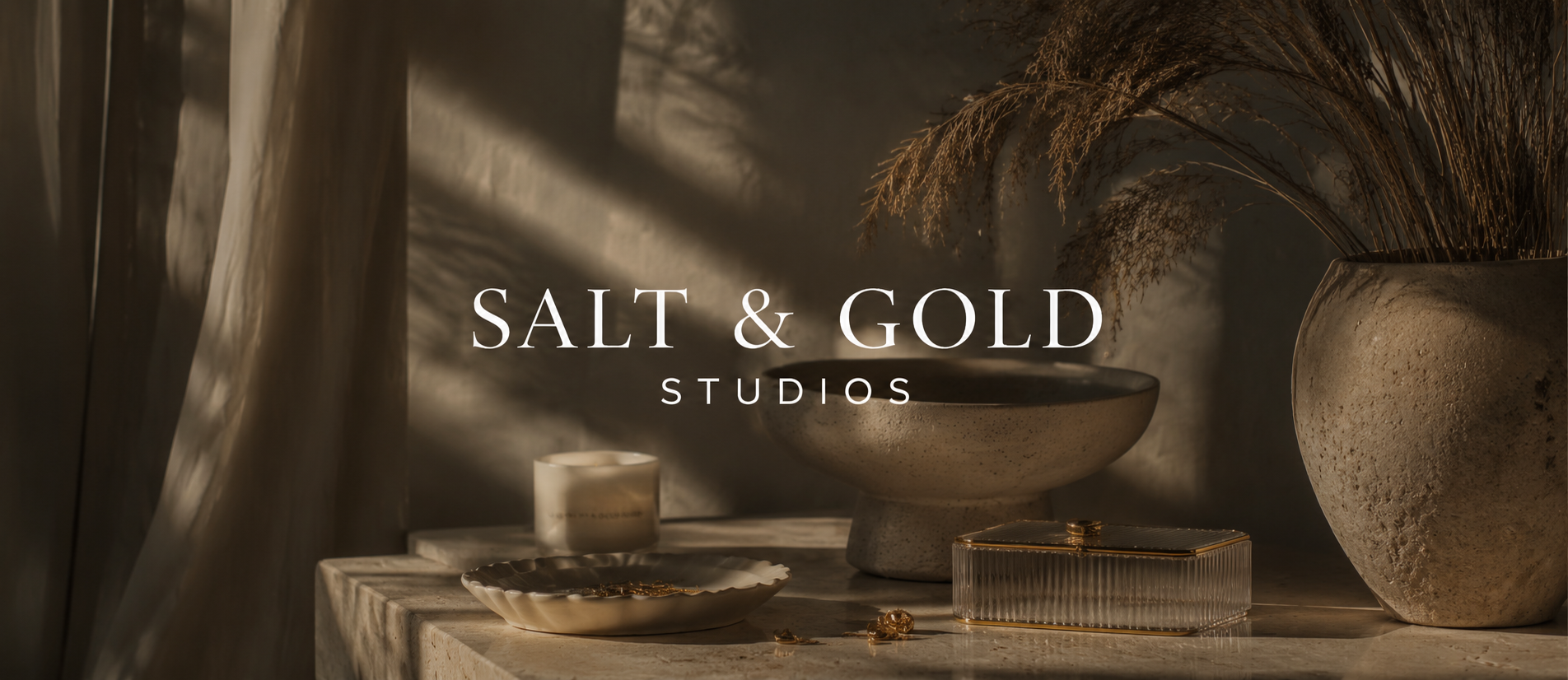 A still life composition featuring a large textured beige vase with dried grasses, a smaller beige bowl, a lit candle, and a rectangular golden jewelry box with earrings, set on a stone surface with dramatic lighting and shadows, with the text "Salt & Gold Studios" overlaid.