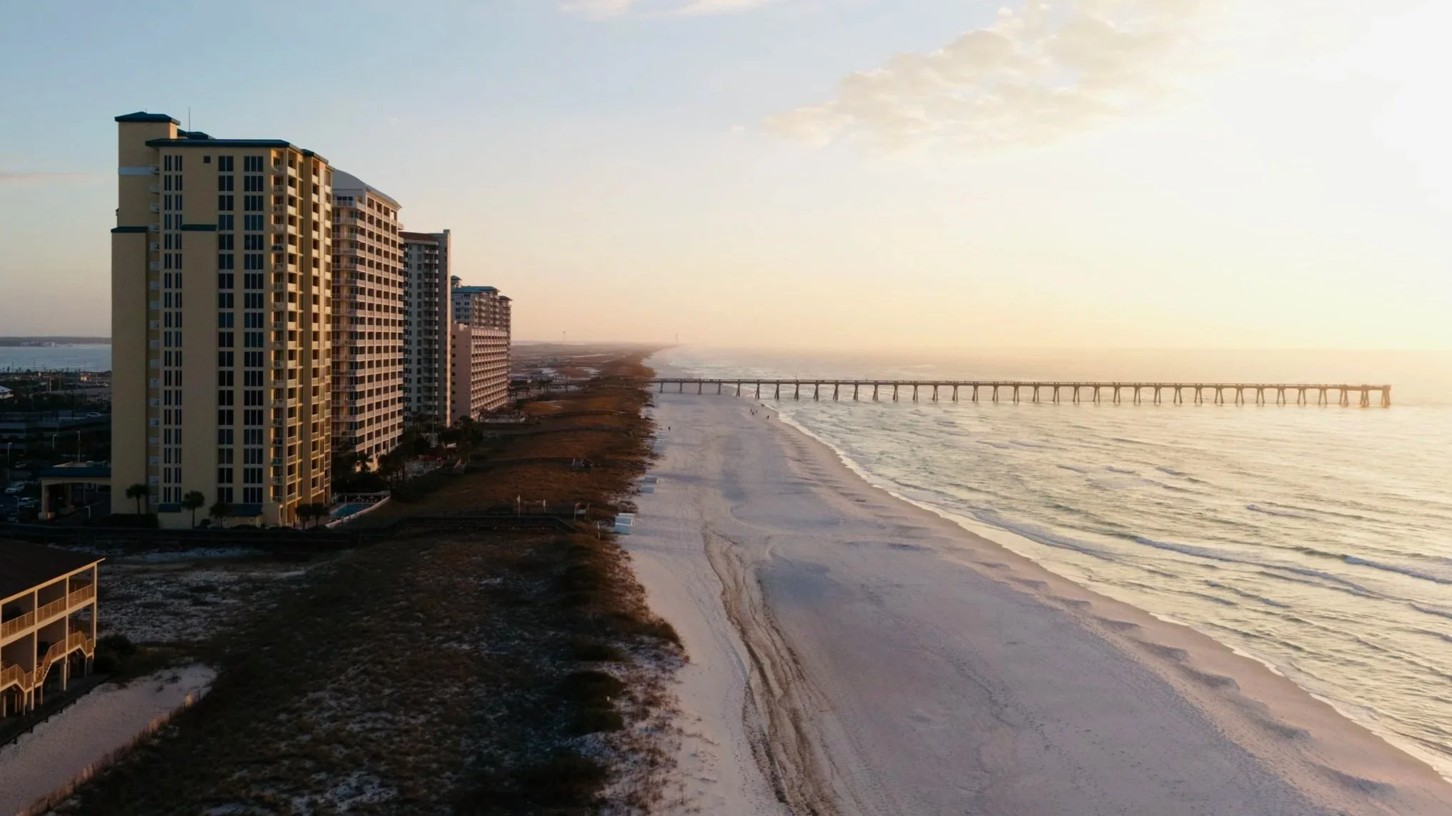 High-rise condominiums along a sandy beach during sunrise, with a long pier extending into the ocean and calm waves.