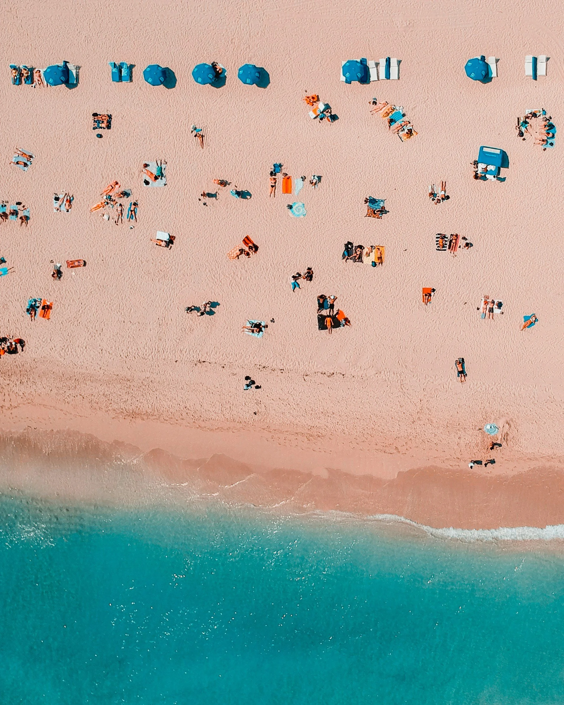 Bird's eye view of a sandy beach with beach umbrellas, lounge chairs, and people relaxing near the ocean.