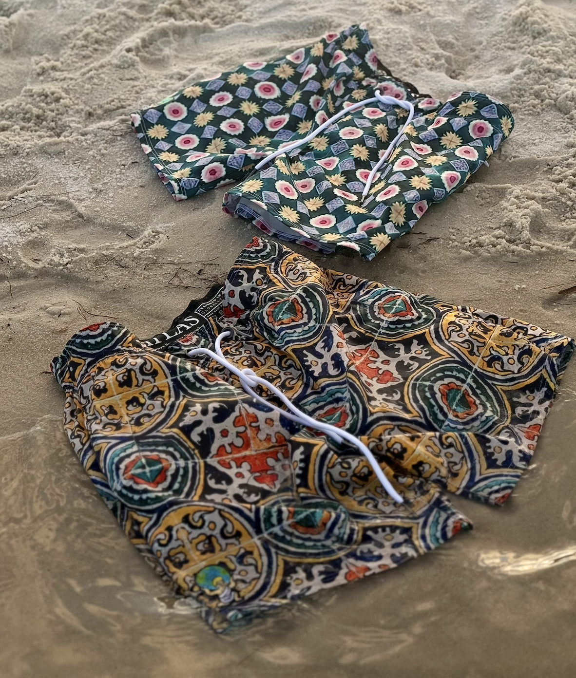 A pair of patterned board shorts on the sand, with a floral swimsuit top nearby.
