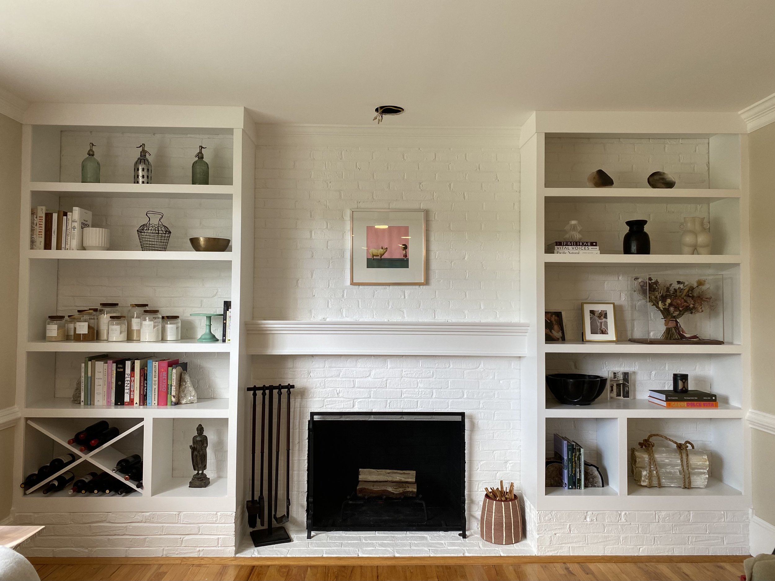 Living room with white brick fireplace, built-in shelves on either side, various decorative items, books, and a small framed painting above the fireplace.