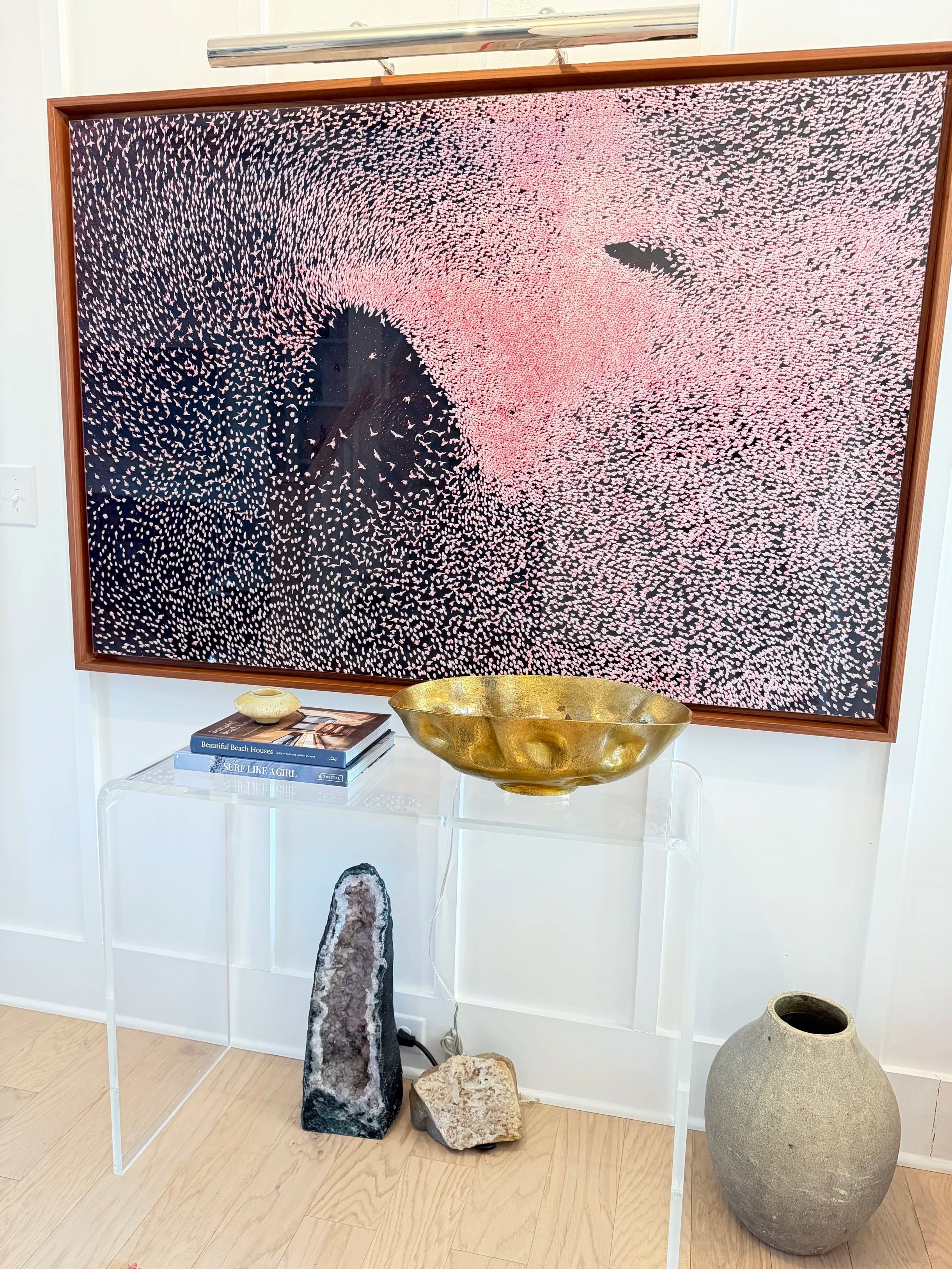 Contemporary art piece with pink, black, and white dots in an abstract pattern, displayed on a wall. In front, a transparent table holds two books, a golden bowl, and a small decorative object. There are two rocks and a large clay pot on the floor be