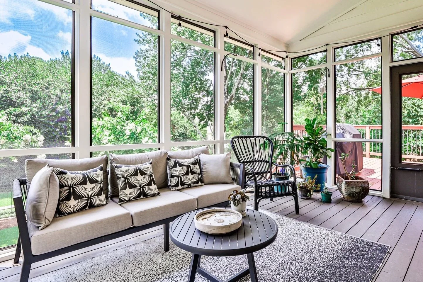 A screened porch with a beige sofa and black-and-white patterned pillows, a black side table, various potted plants, and a wooden deck with a red umbrella visible outside.