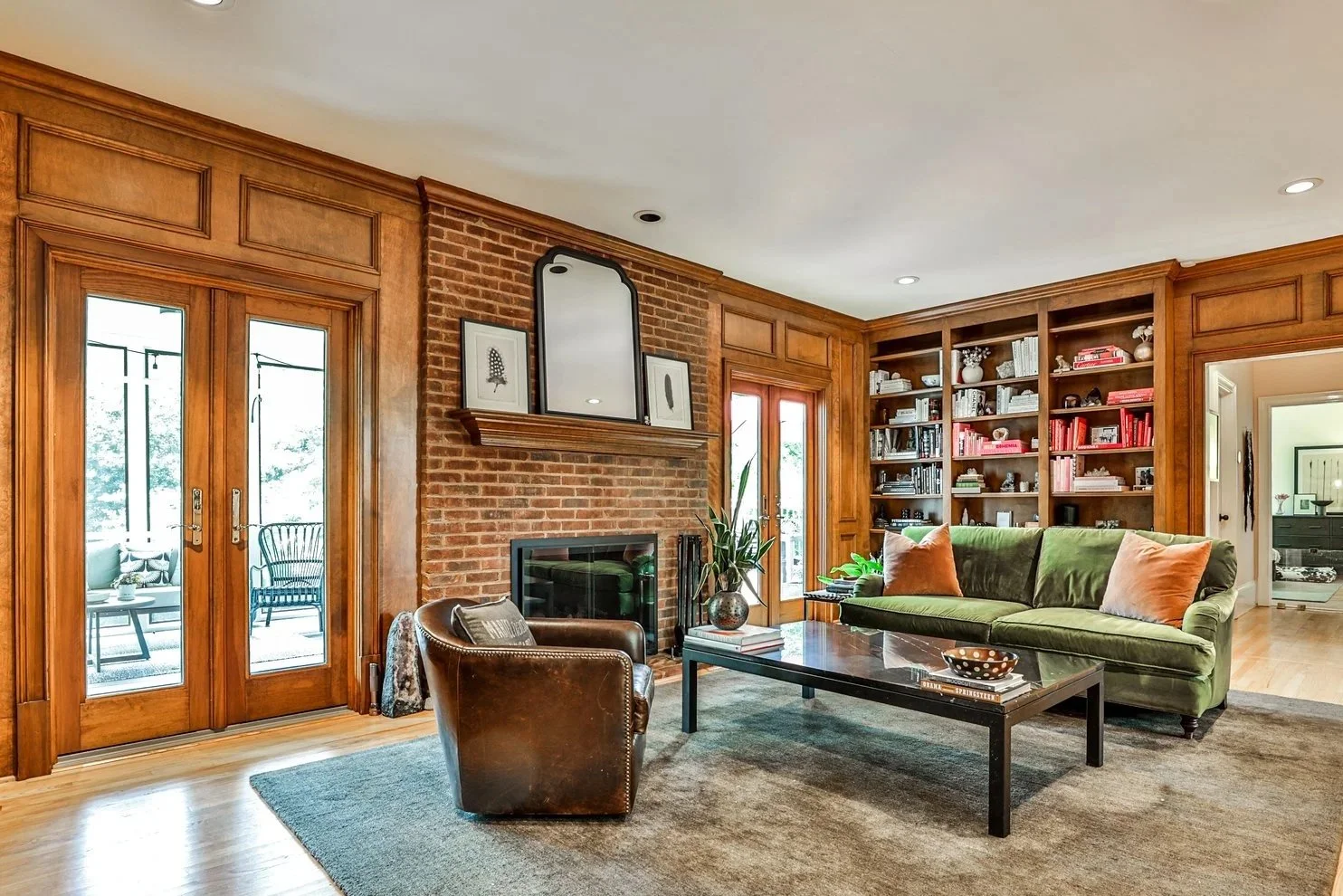 Living room with brick fireplace, wooden built-in shelves filled with books and decor, green sofa with orange pillows, leather armchair, glass coffee table, sliding glass doors leading outside, and cozy decor.