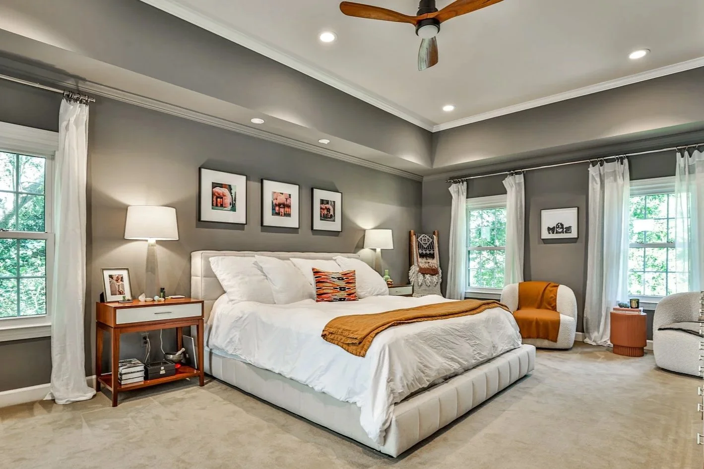 A stylish bedroom with gray walls, a large white bed with white bedding and decorative pillows, wooden nightstands with lamps, framed artwork above the bed, windows with white curtains, and cozy seating with orange and beige accents.