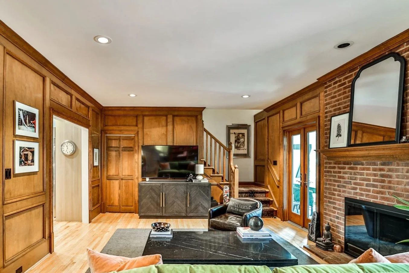 Living room with wood-paneled walls, a brick fireplace with a mirror above, a large TV on a cabinet, a black coffee table with books and decor, a leather armchair, and a wooden staircase with a glass door leading outside.