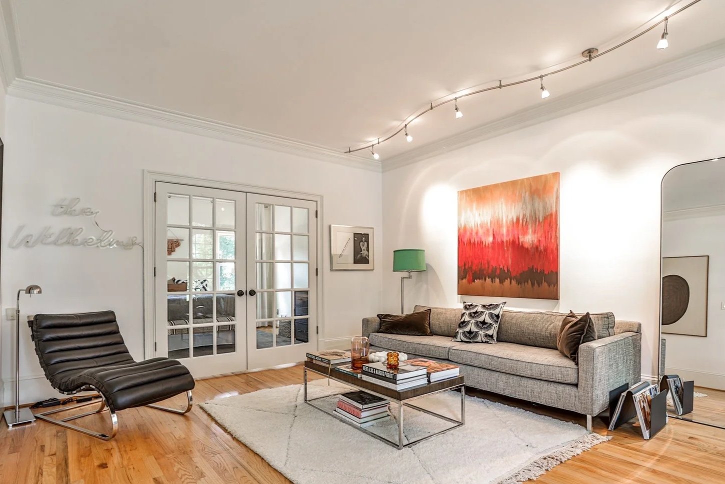 Living room with beige sofa, colorful abstract wall art, black lounge chair, glass doors, green floor lamp, white walls, hardwood floor, decorative items on coffee table, wall sign reading 'cheer regardless'.