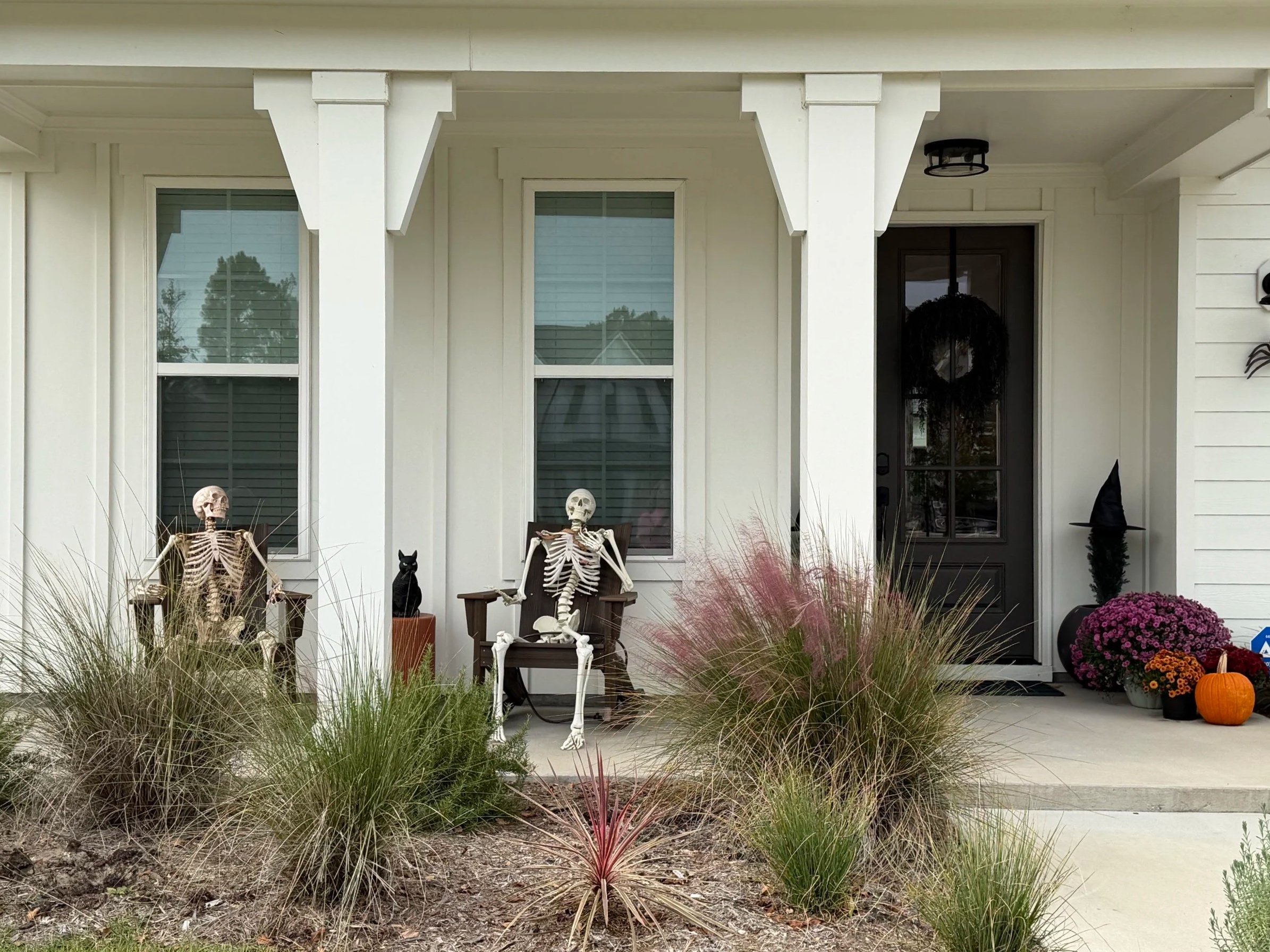 Front porch decorated for Halloween with two skeletons seated in chairs, black cat figurine, pumpkin, purple chrysanthemums, and witch hat.