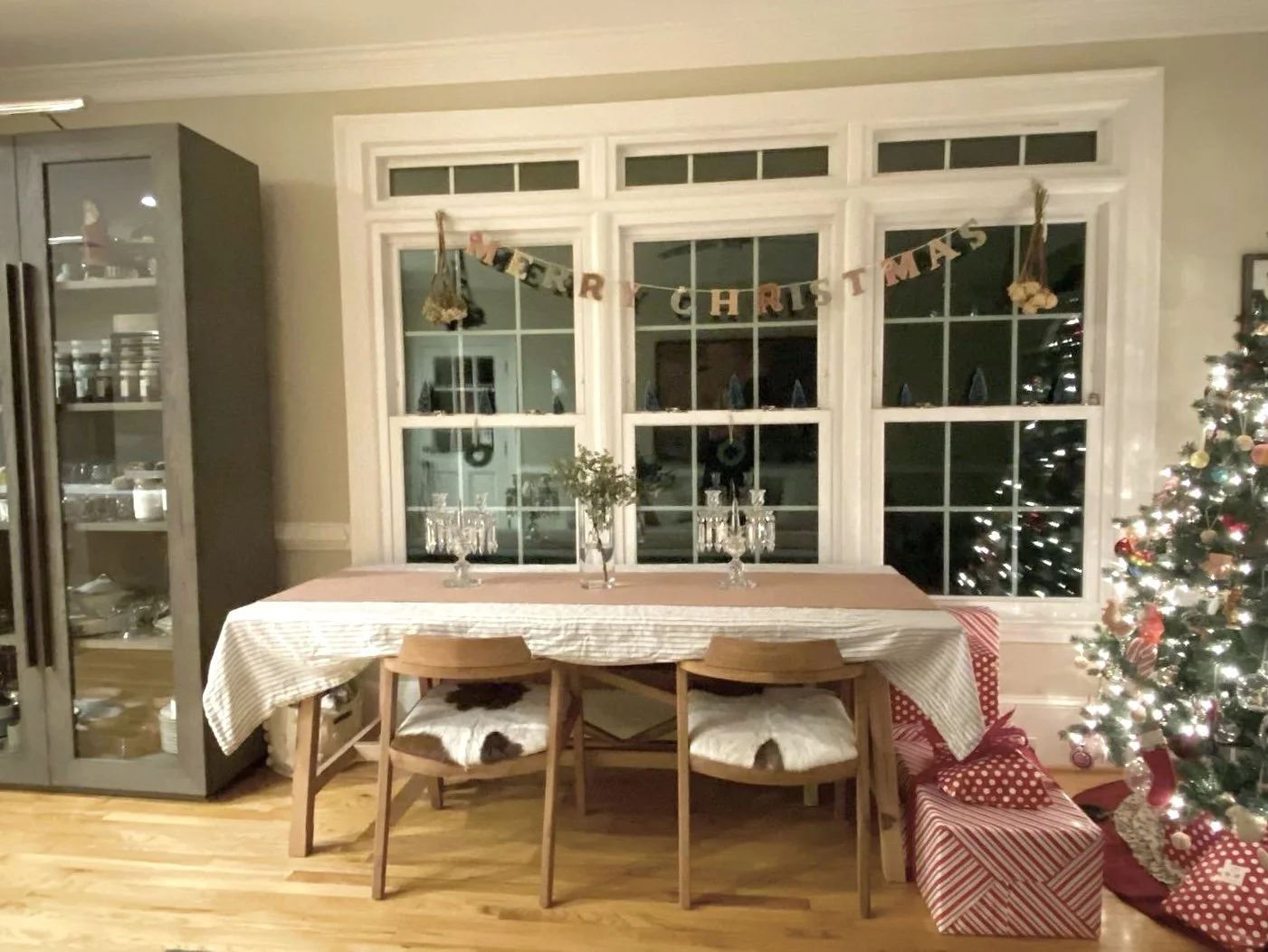 Dining room decorated for Christmas with a festive banner, a Christmas tree with ornaments and lights, a table with a tablecloth, candles, and a vase with greenery, and wrapped presents under the tree.