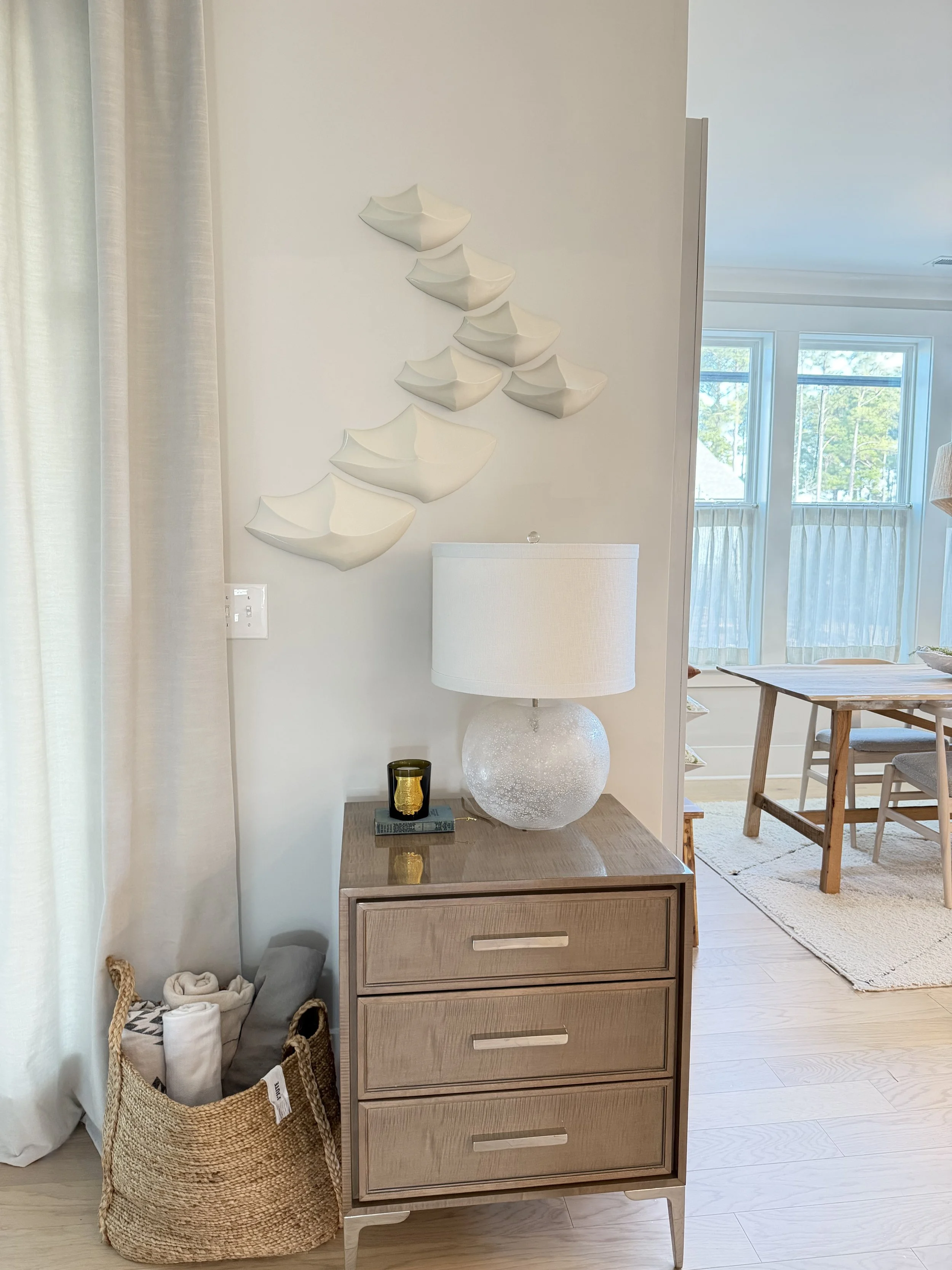 A wooden bedside table with a textured white lamp and small decor items, beside a woven basket filled with rolled towels, in a modern bright room with large windows and white curtains, decorated with white wall art resembling boats.