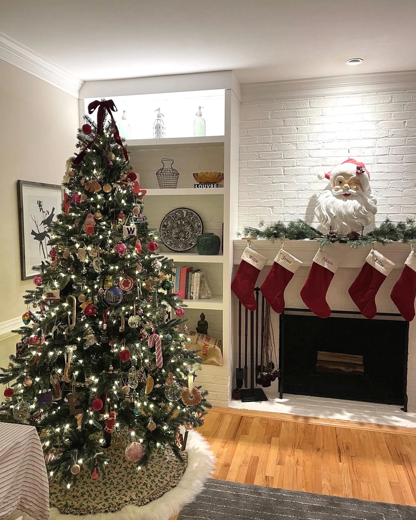 Decorated Christmas tree with lights, ornaments, and a tree skirt, positioned next to a white shelf with decorative items, and a white brick fireplace mantel adorned with holiday stockings and a Santa head decoration.