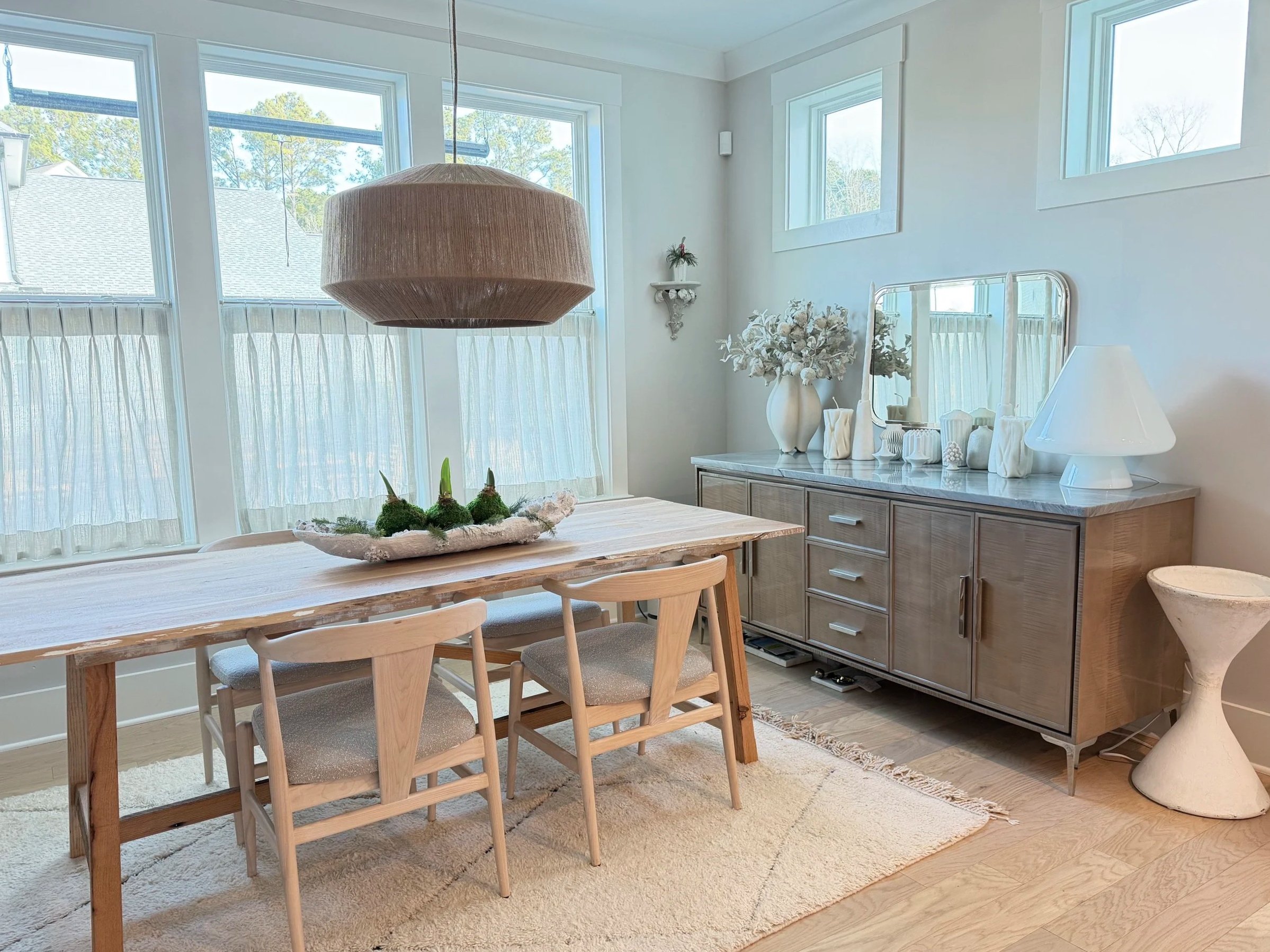 Bright dining room with large windows and sheer curtains, wooden table with three chairs, modern sideboard with decorative vases, mirror, and a white table lamp, and a large pendant light hanging above the table.