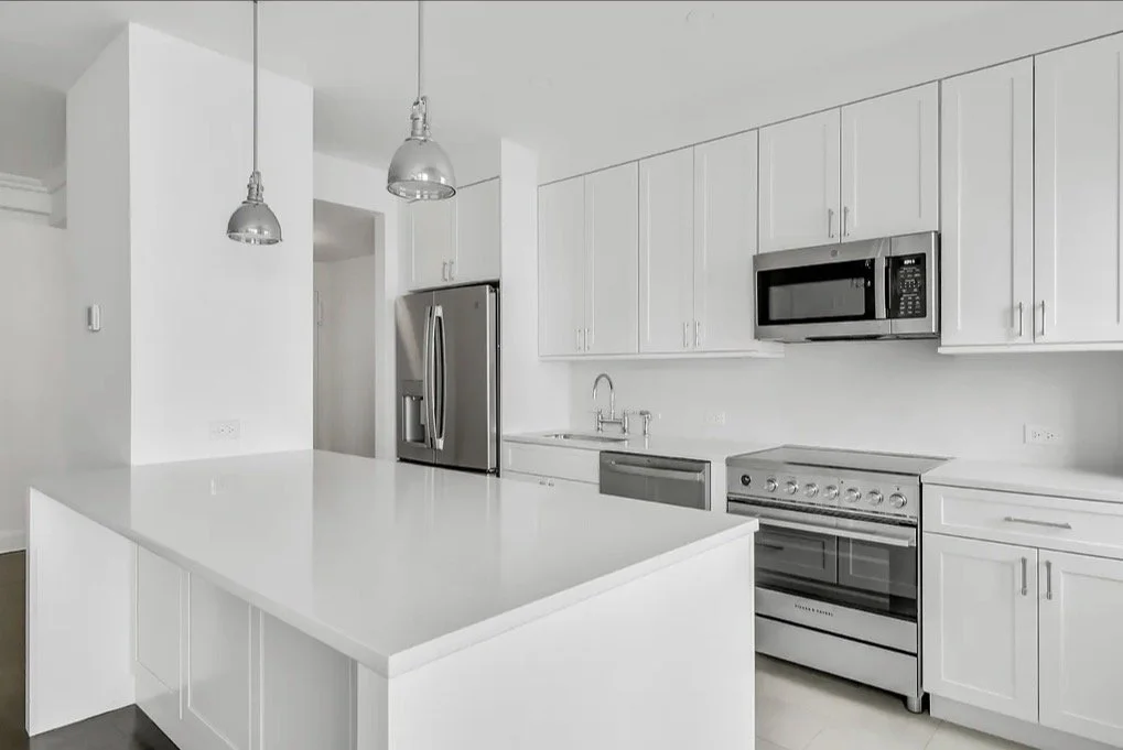 Modern white kitchen with stainless steel appliances, including refrigerator, microwave, and oven, and two pendant lights hanging over a kitchen island.