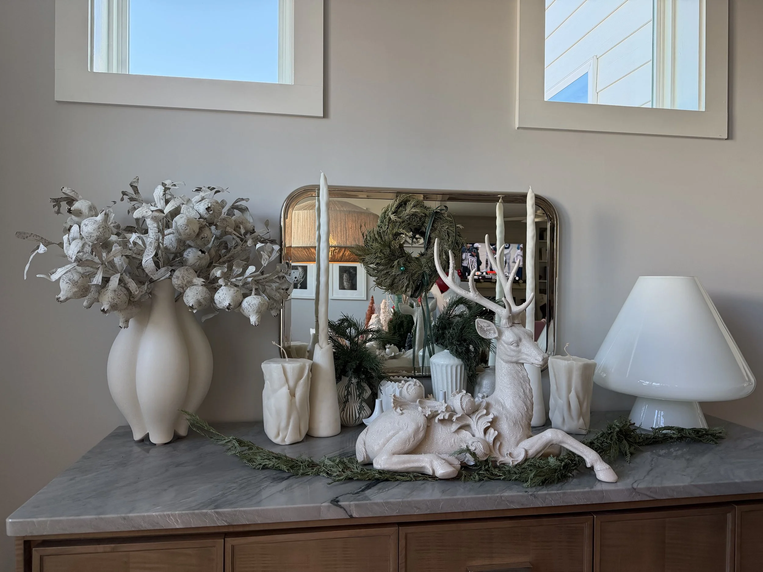 Decorative holiday display on a gray marble-top sideboard, featuring white vases with cotton branches, a white ceramic deer figurine, candles, yellowed greenery, and a wreath reflected in the mirror, with two windows above.
