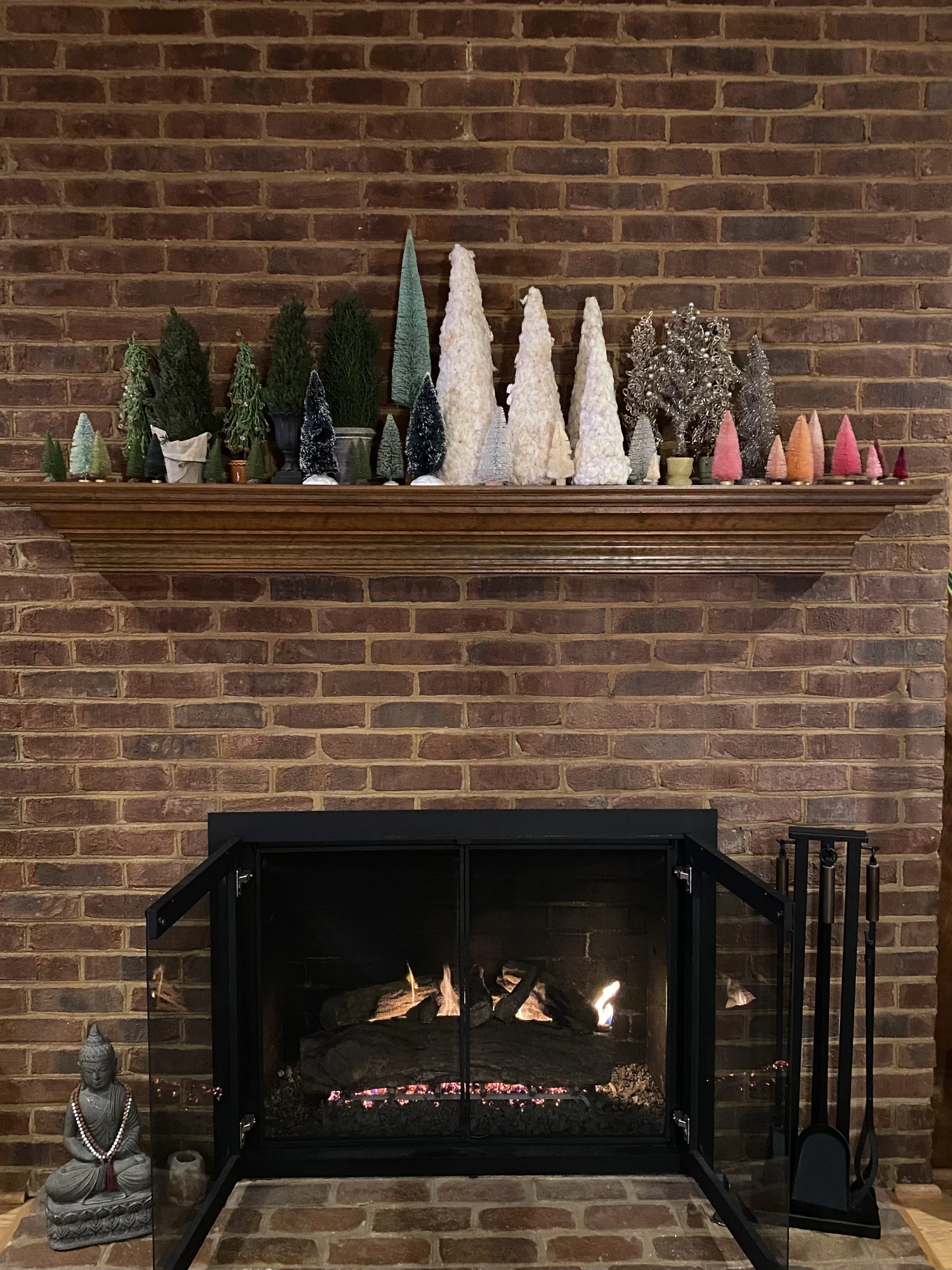 Decorative Christmas trees on a wooden mantel above a lit fireplace, with a Buddha statue and a small jar or candle on the hearth.