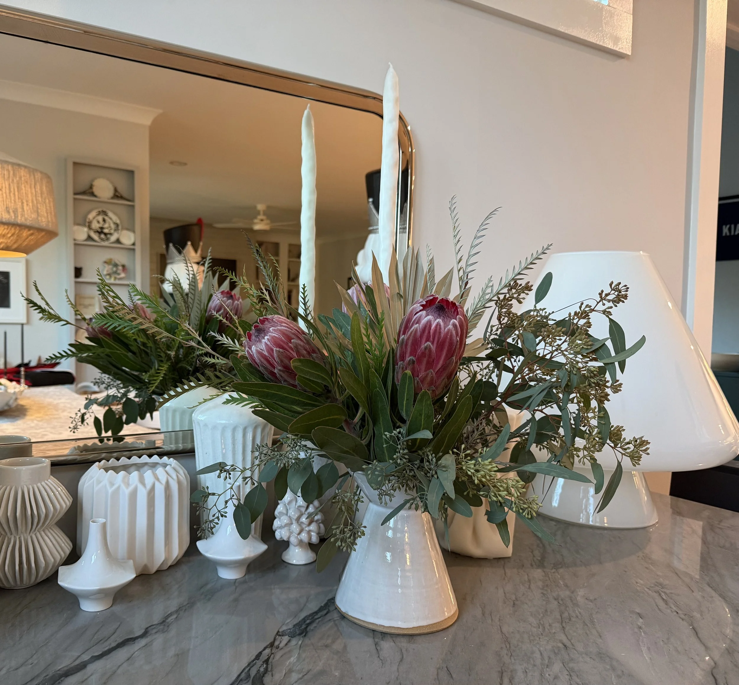 A floral arrangement with protea flowers, greenery, and candles in white ceramic vases on a gray marble surface, with a mirror and a white lamp in the background.