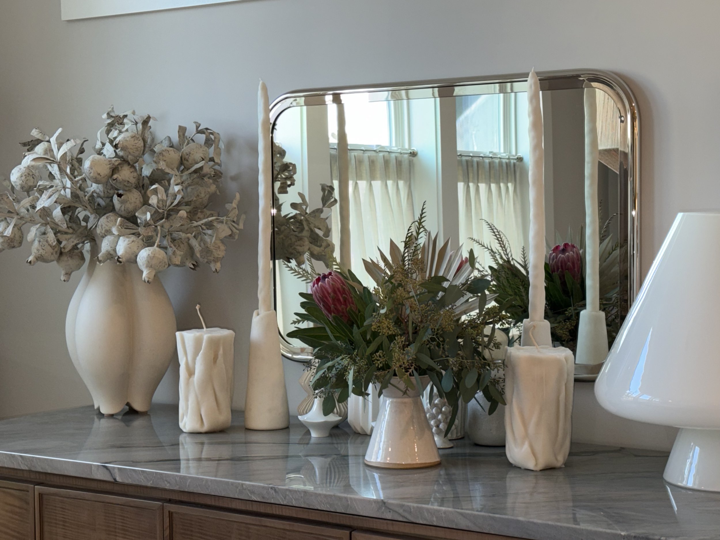 Decorative vases with dried and fresh flowers and candles, placed on a marble tabletop in front of a mirror.