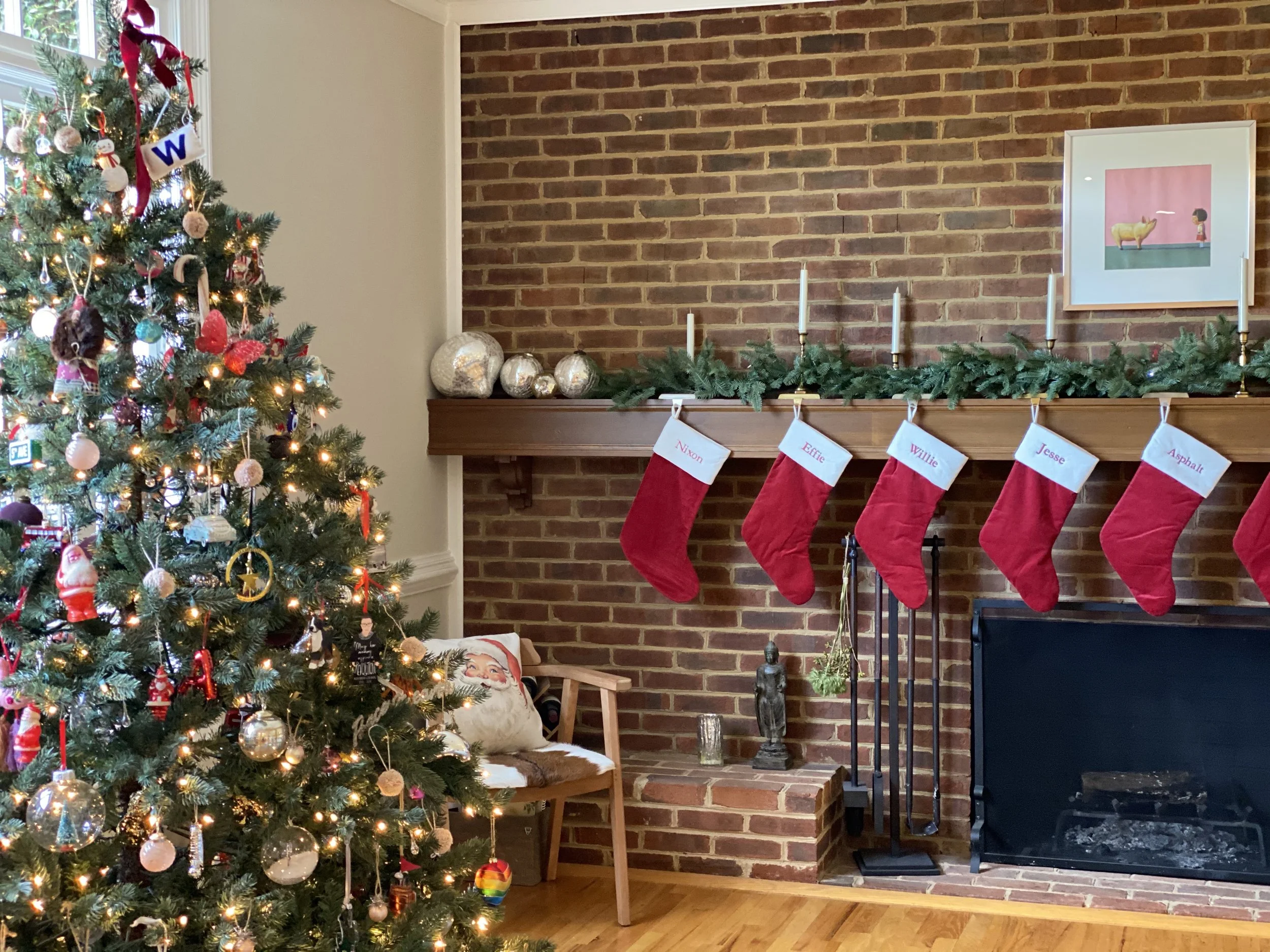 A decorated Christmas tree with ornaments and lights beside a brick fireplace with stockings hanging. The mantel has greenery and candles, with a framed picture of a girl and a goat above.