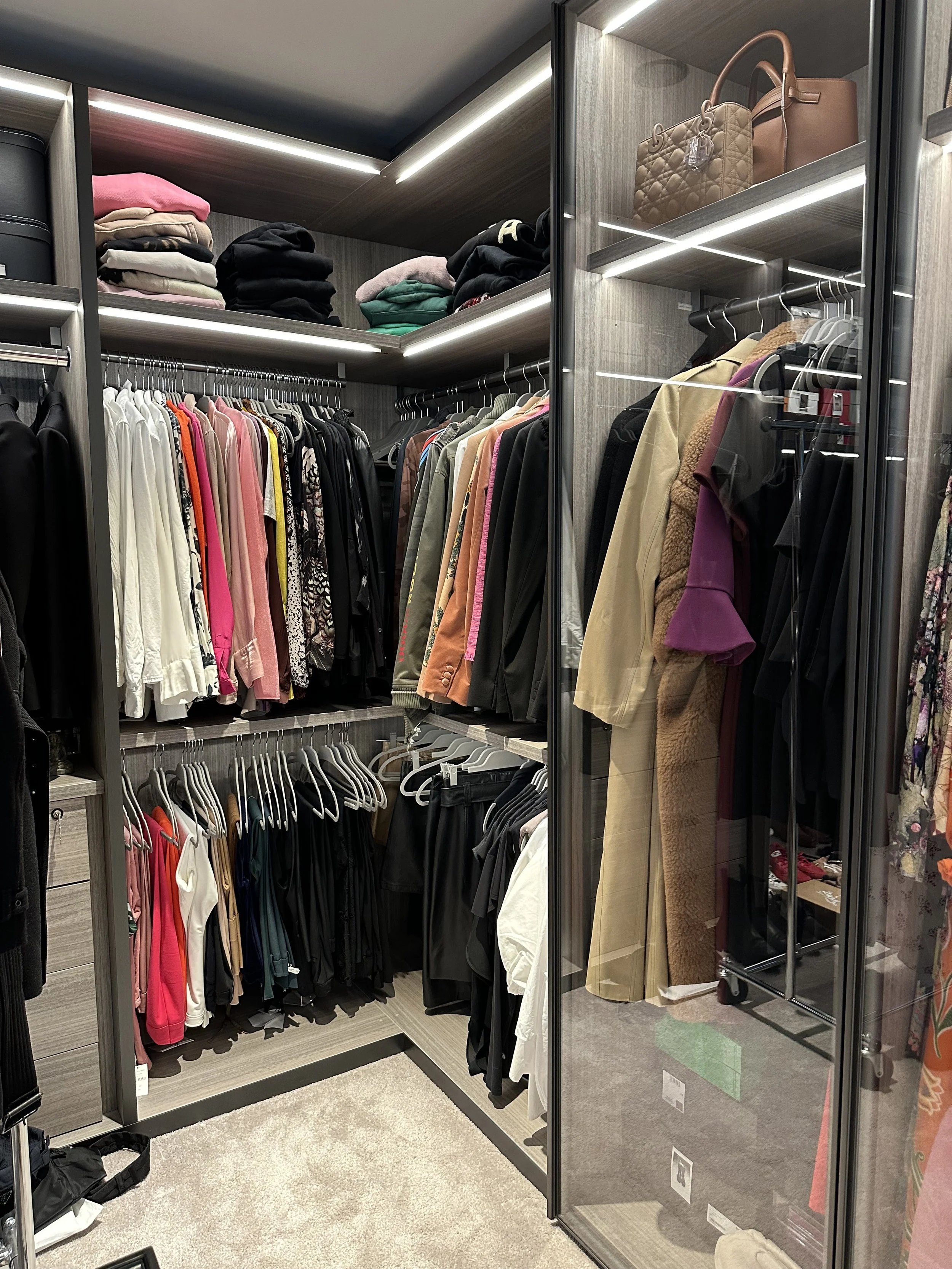 A walk-in closet with shelves full of folded clothes, hanging shirts, jackets, and pants, along with some handbags and accessories on the upper shelves.