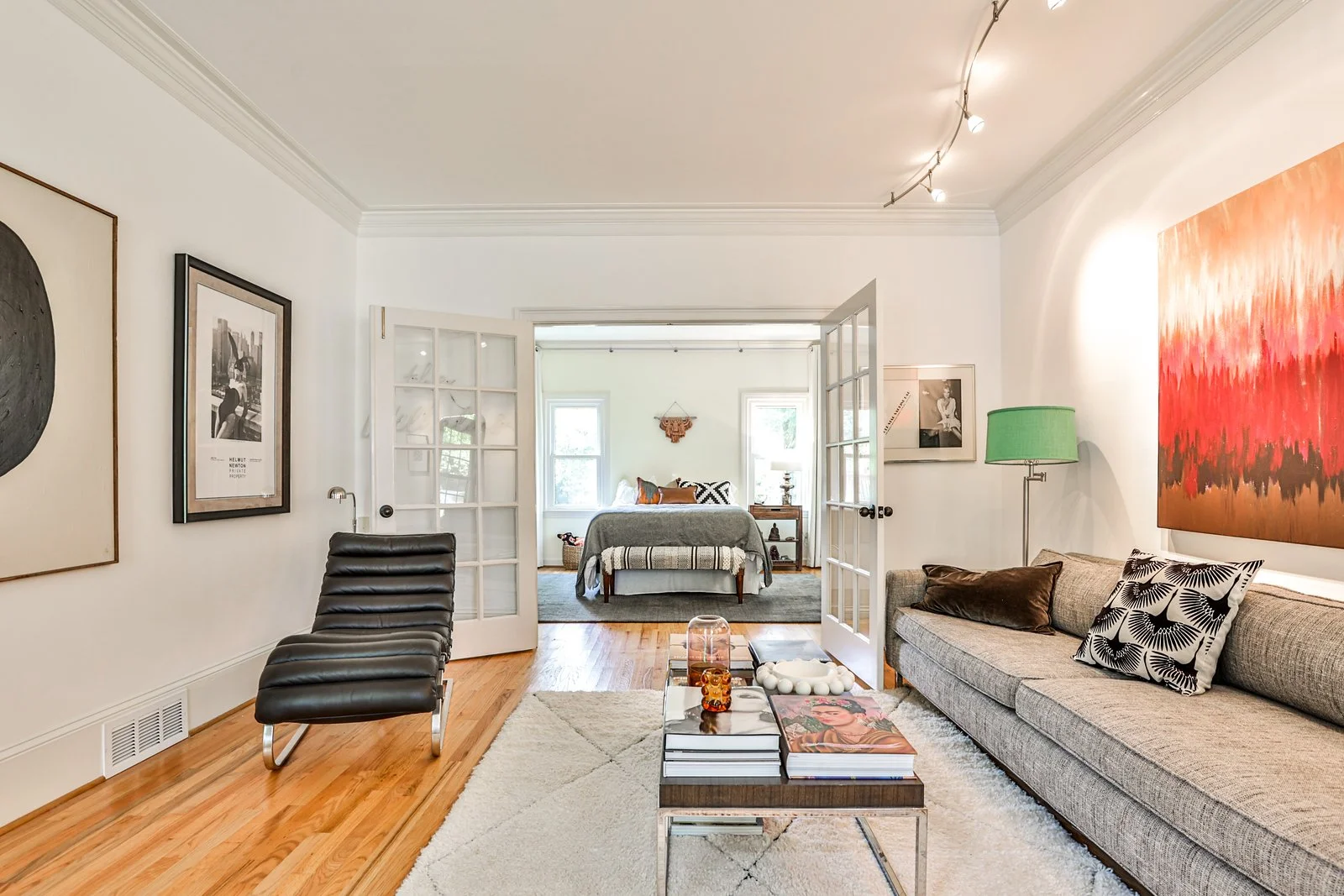 Living room with beige sofa, colorful artwork, black chair, coffee table with books and decor, and an adjoining bedroom with a bed, window, and wall art.