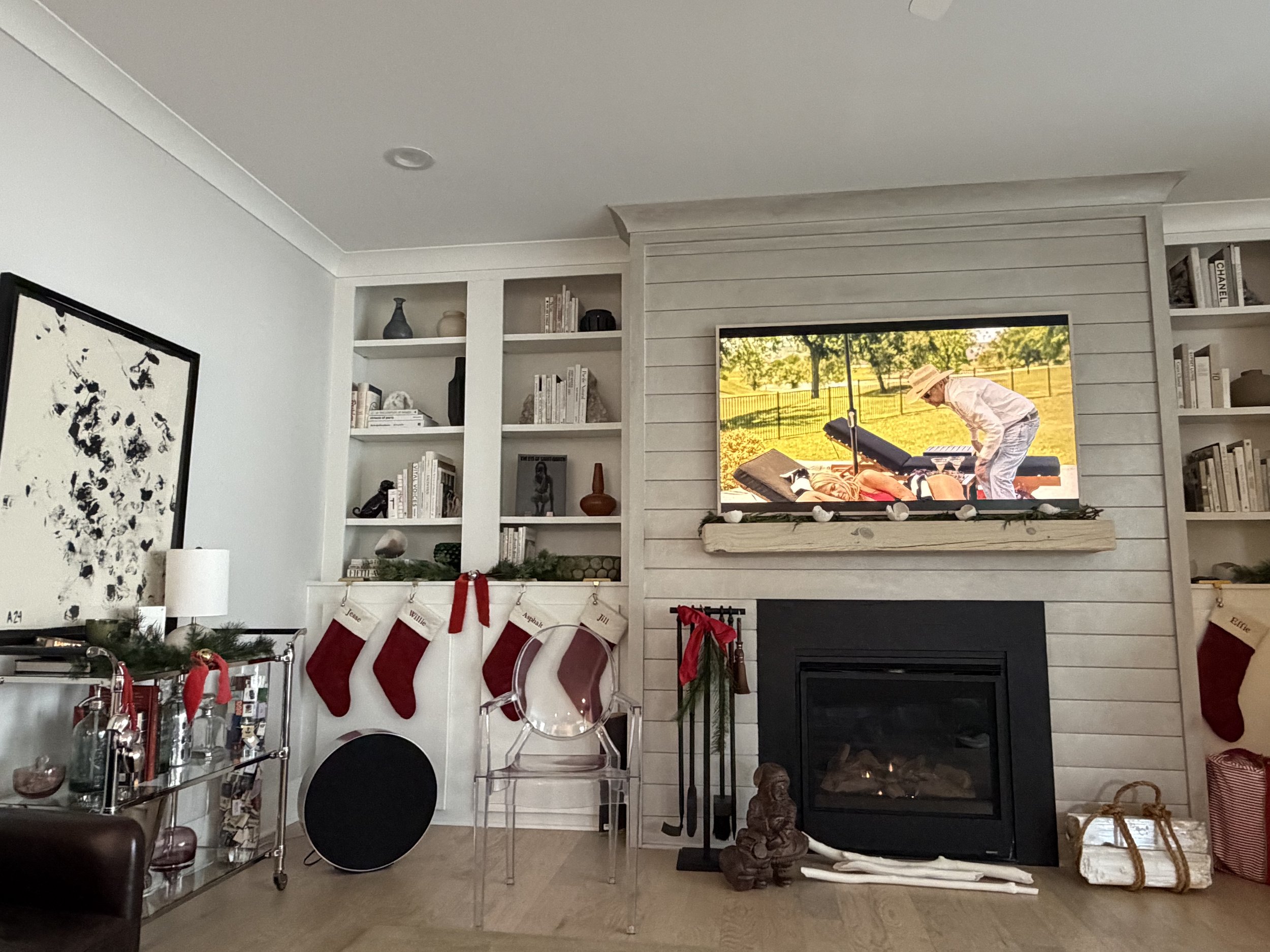 Living room decorated for Christmas with stockings hanging on the fireplace, a Christmas wreath on the side table, and stockings hung on the wall above the fireplace. A flat-screen TV on the wall displays a scene with a person in a hat leaning over a