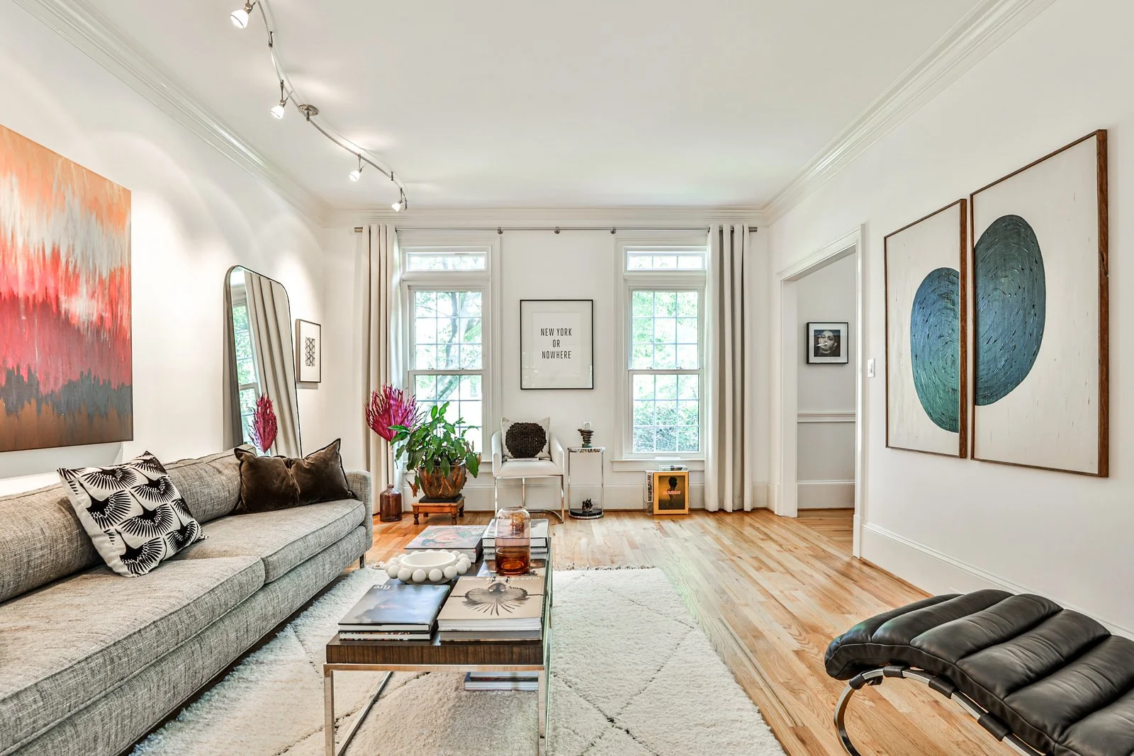A living room with hardwood floors, white walls, and large windows. There is a beige sofa with decorative pillows, a coffee table with books and decor, a black lounge chair, and wall art including abstract paintings. A potted plant with pink flowers 