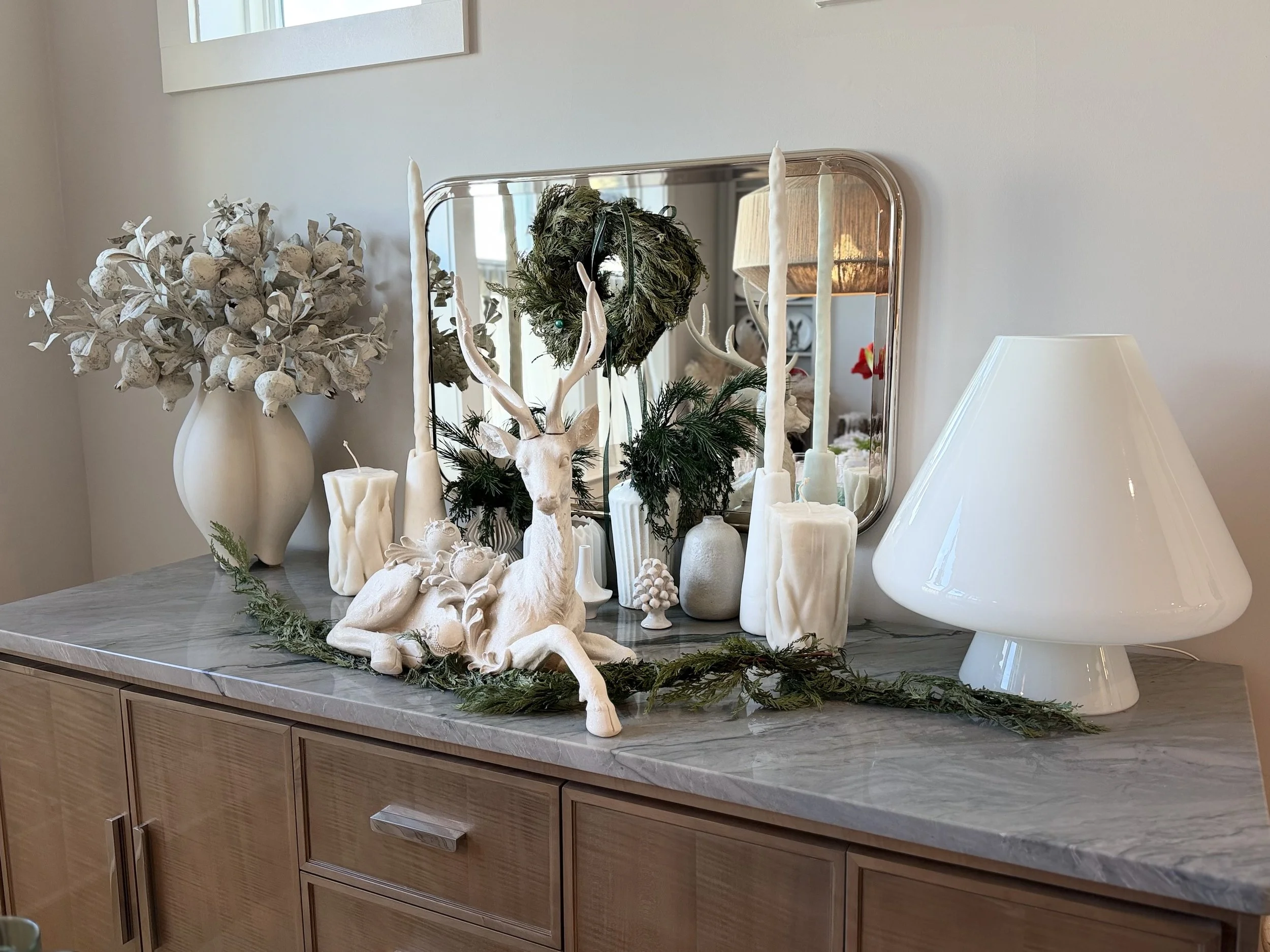 Decorative holiday display on a wooden sideboard featuring a white reindeer figurine, white candles, greenery, a white ceramic vase with dried flowers, a large white table lamp, and a mirror reflected above.