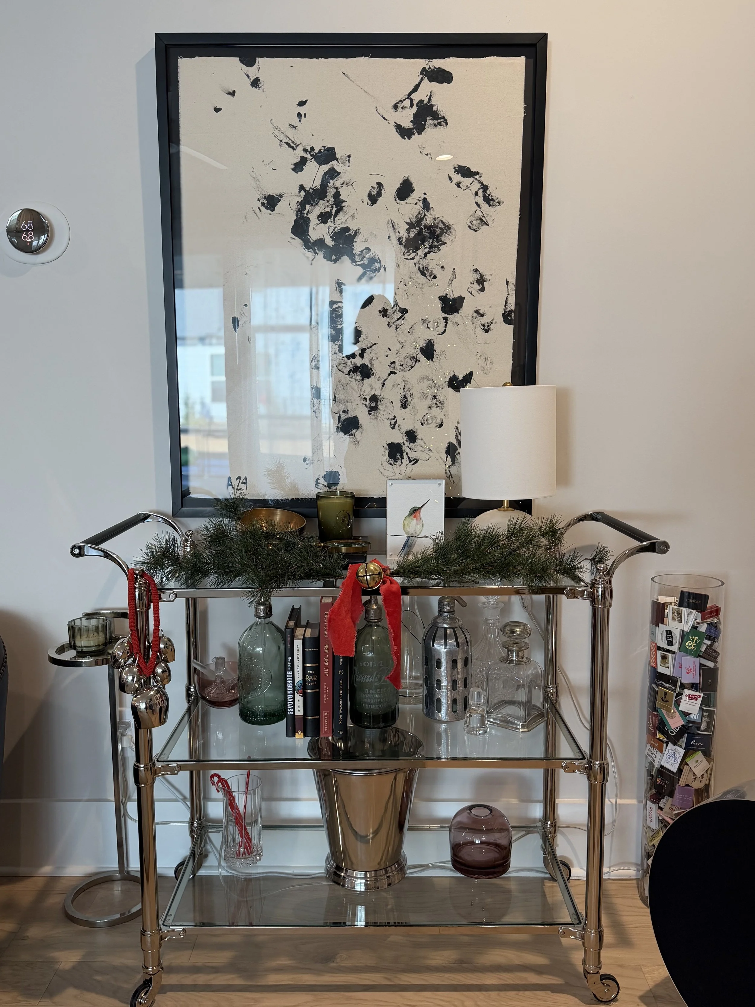 Decorative bar cart with glass bottles, books, and holiday ornaments, topped with greenery, against a wall with abstract black and white artwork.