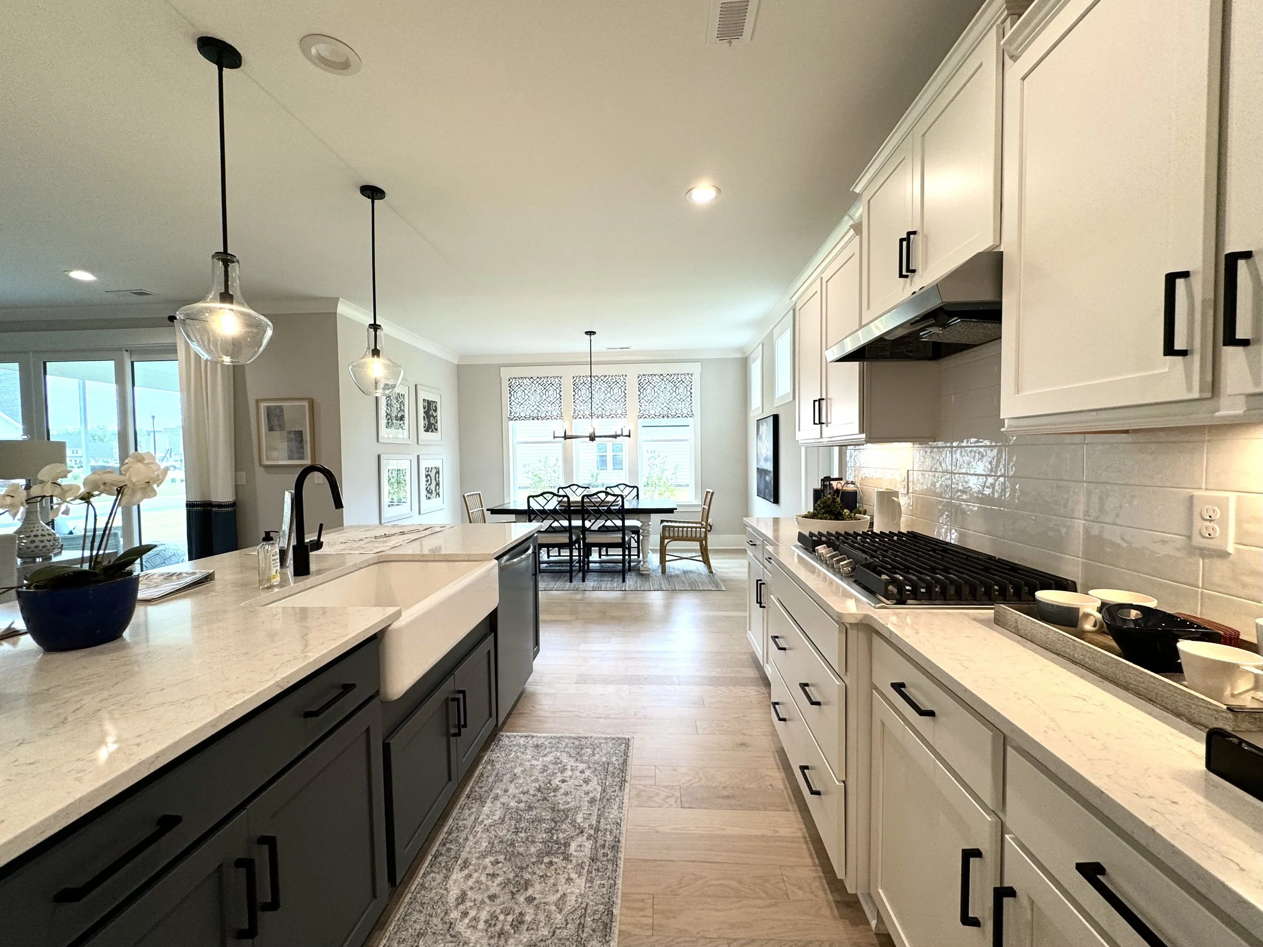 Modern kitchen with white cabinets, black hardware, white countertops, a large island with a farmhouse sink, pendant lights, and a view of the dining area with large windows and a patio door.