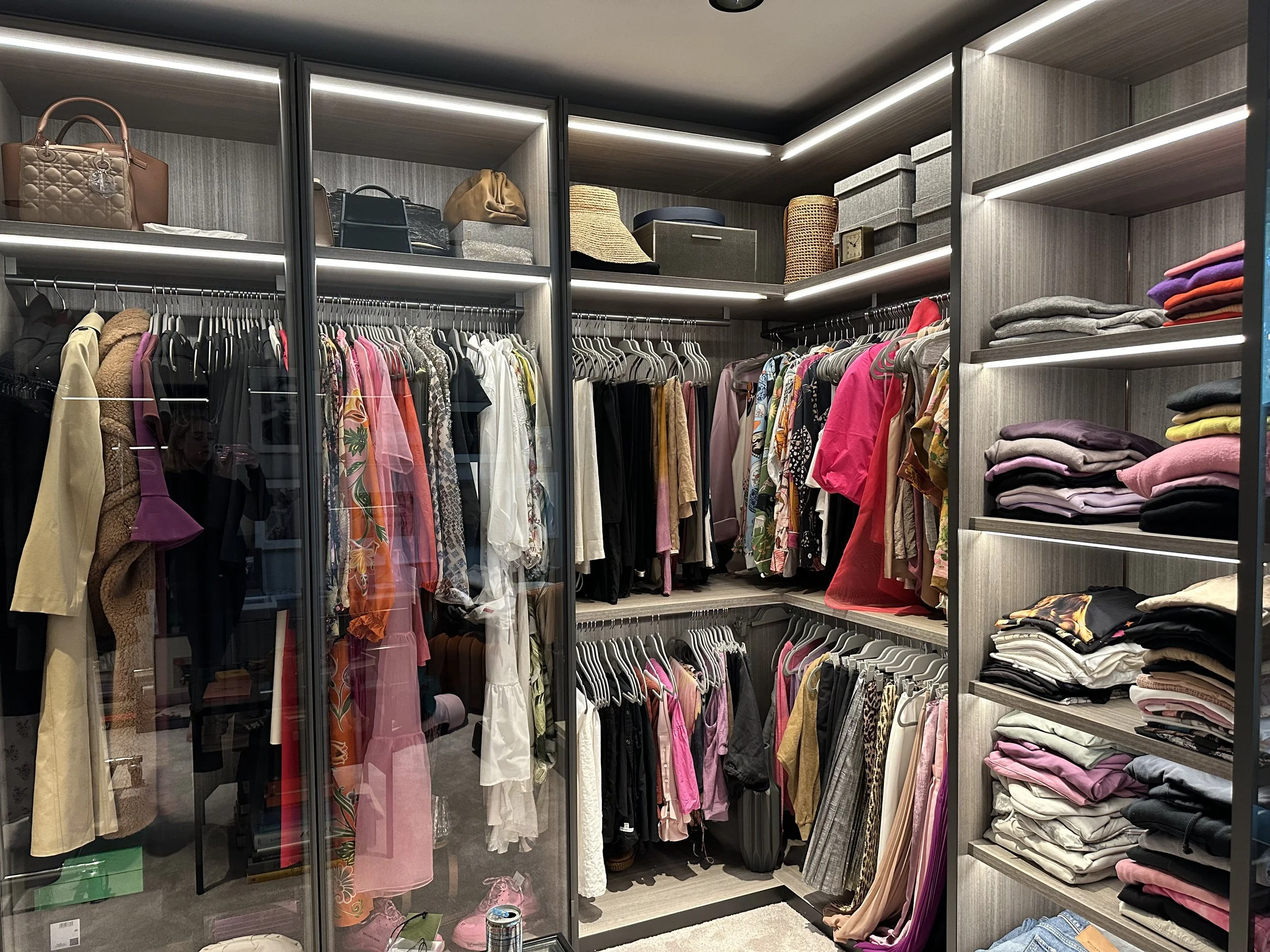 A walk-in closet with hanging clothes, folded sweaters on shelves, and handbags on the top shelf.