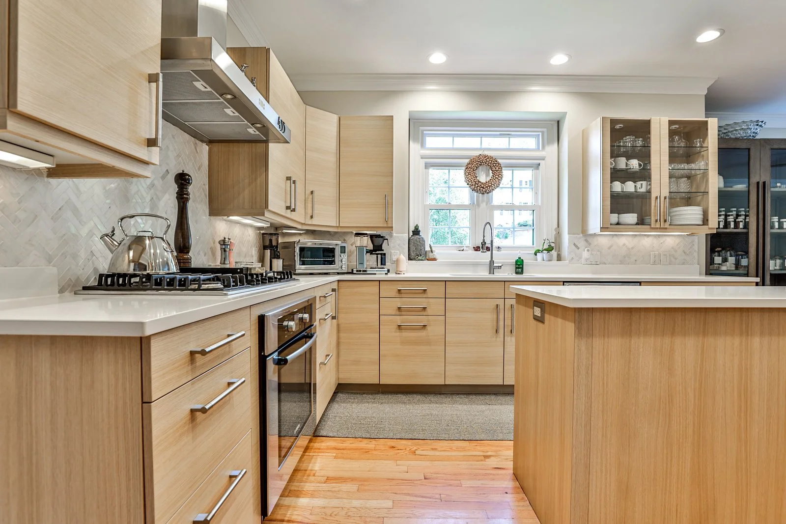 A modern kitchen with light wood cabinets, white countertops, stainless steel appliances, and hardwood floor. A window with a wreath above the sink lets in natural light.