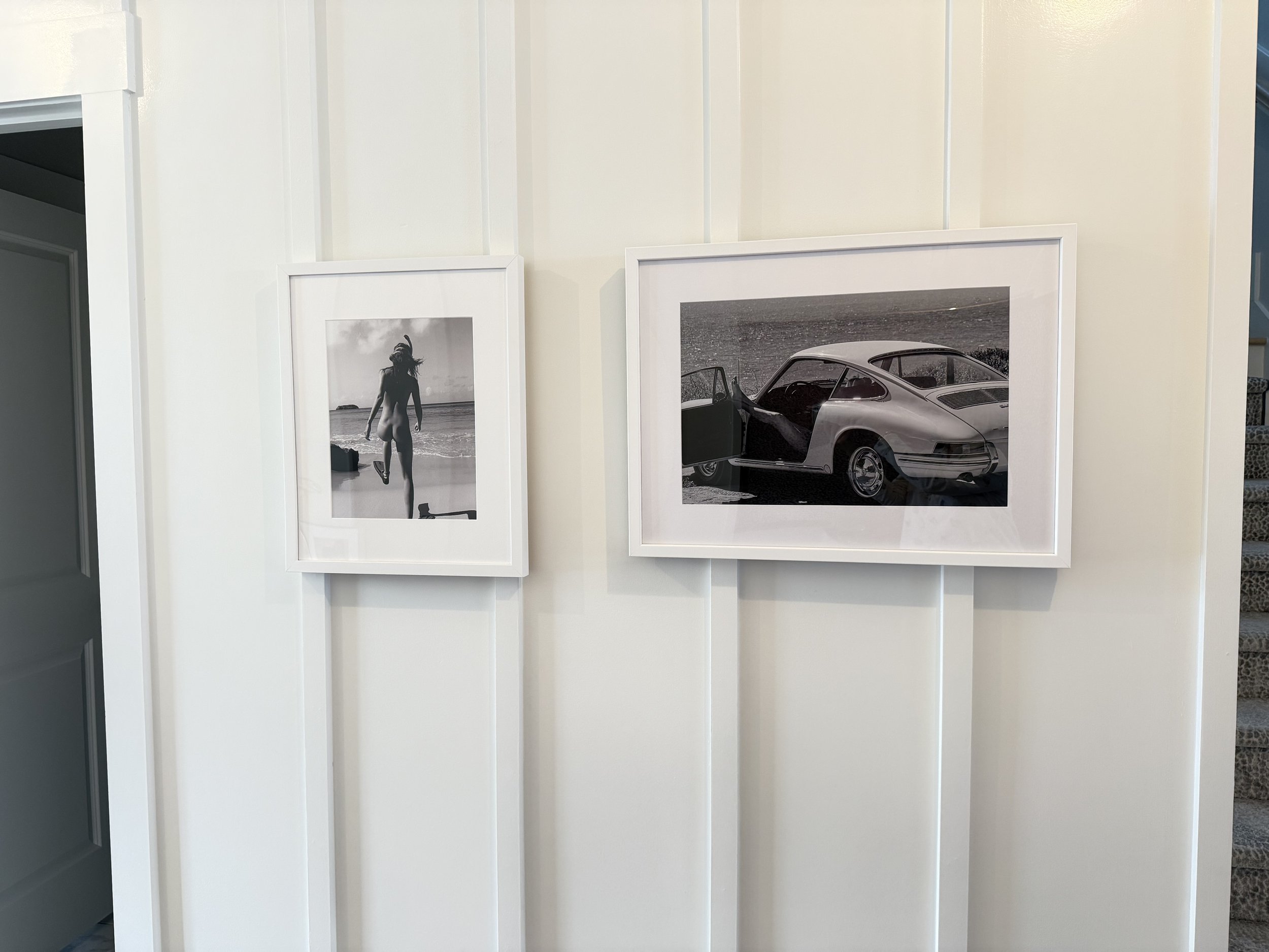 Black and white photographs on white wall, one showing a woman walking on the beach, the other showing a vintage car with open door.