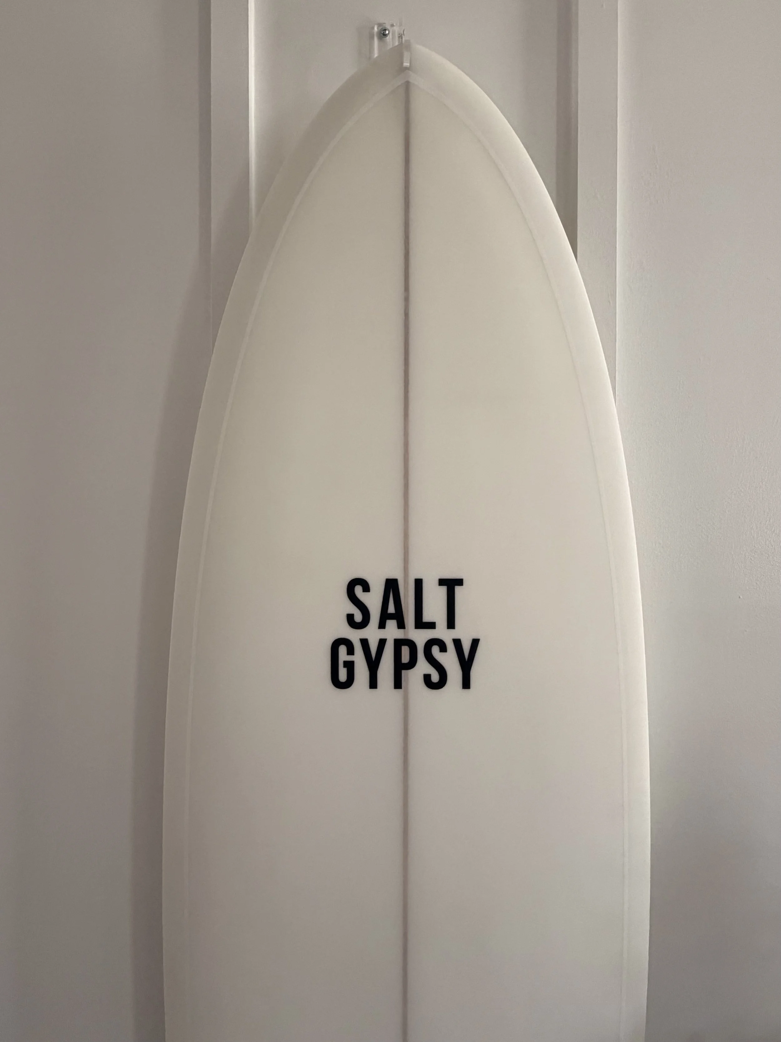 A white surfboard with the words "SALT GYPSY" in black letters, leaning against a wall.