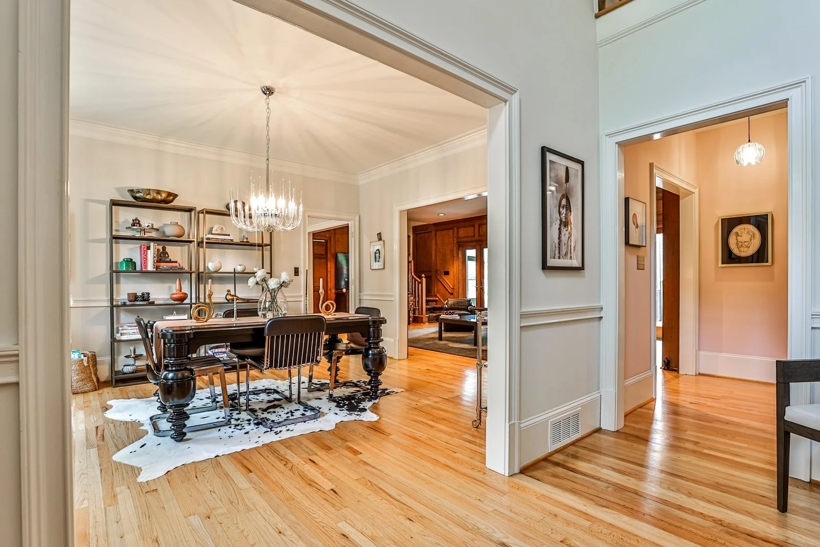 Dining room with a black dining table, four chairs, a metal bookshelf, a modern chandelier, a cowhide rug, and a wooden floor.
