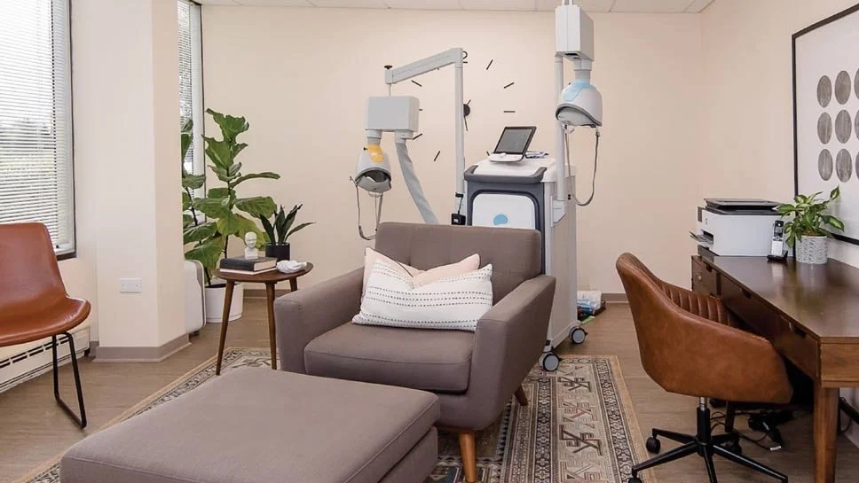 Medical office room with a brown chair, gray armchair with a pillow, small table with plant, and medical equipment in the background.