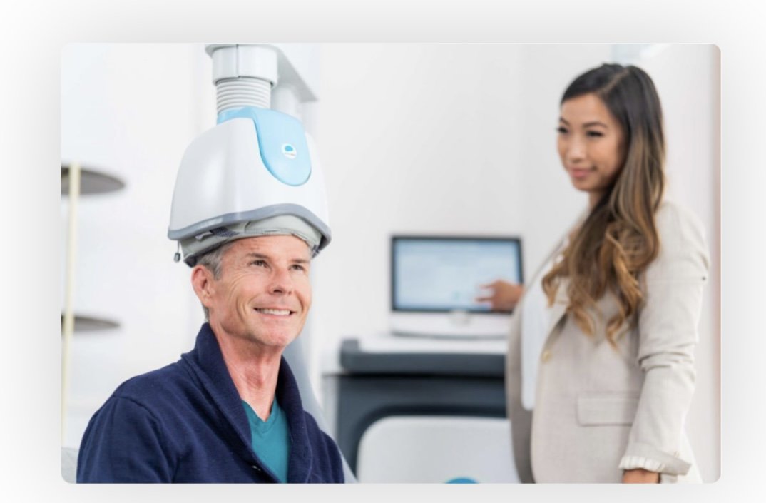 A man undergoing a medical scan with a machine on his head, in a clinical setting with a woman staff member standing nearby.