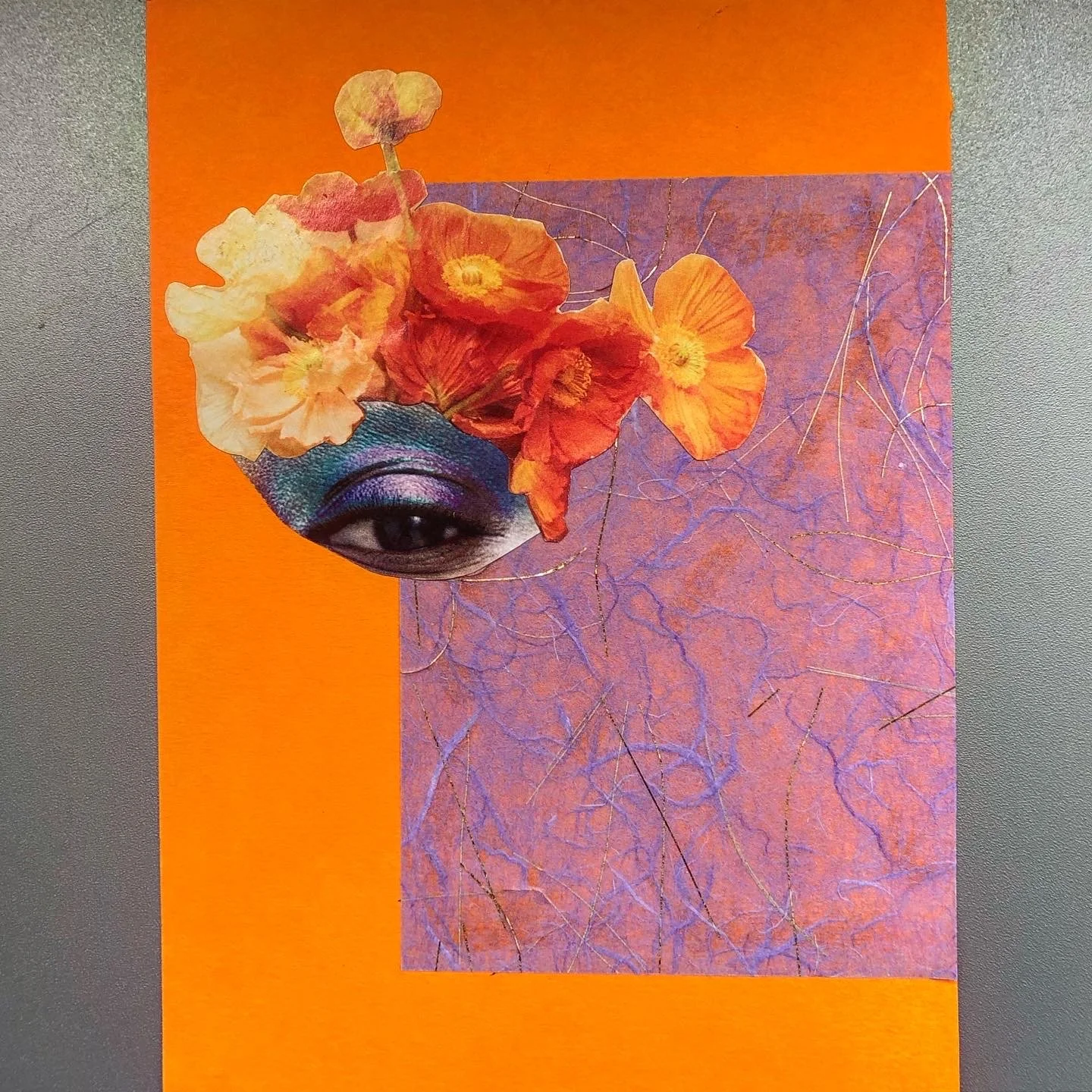 A collage featuring an eye with purple and blue eyeshadow, orange and yellow flowers around eye brow, and a background of purple handmade japanese paper with gold and purple fibres on an orange card background. on grey surface