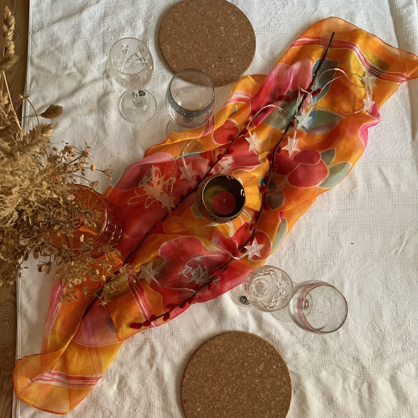 A table setting with a white tablecloth, decorative orange, pink, and green floral fabric centerpiece, string lights, wine glasses, water glasses, a small candle in a dark holder, two round cork placemats, and dried flowers in a vase.