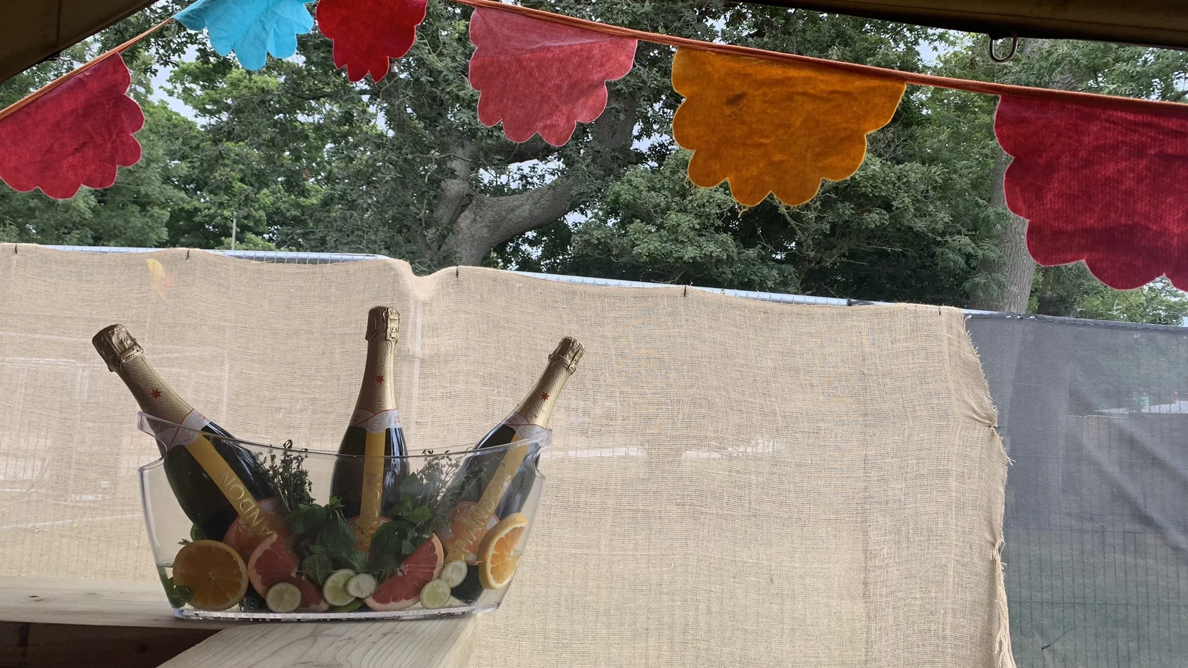 Three bottles of champagne with gold foil on their necks are placed in a clear bowl filled with fresh citrus slices and herbs for a brand showcase. The outdoor festival bar has colorful paper decorations hanging from the tent.
