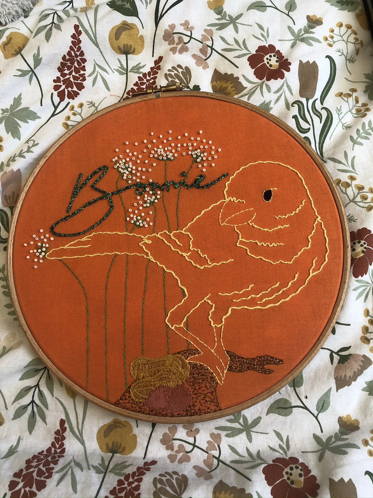 Embroidery artwork of a dinosaur and flowers on an orange fabric background, with the name 'Bambie' stitched above the flowers, surrounded by a floral patterned surface.