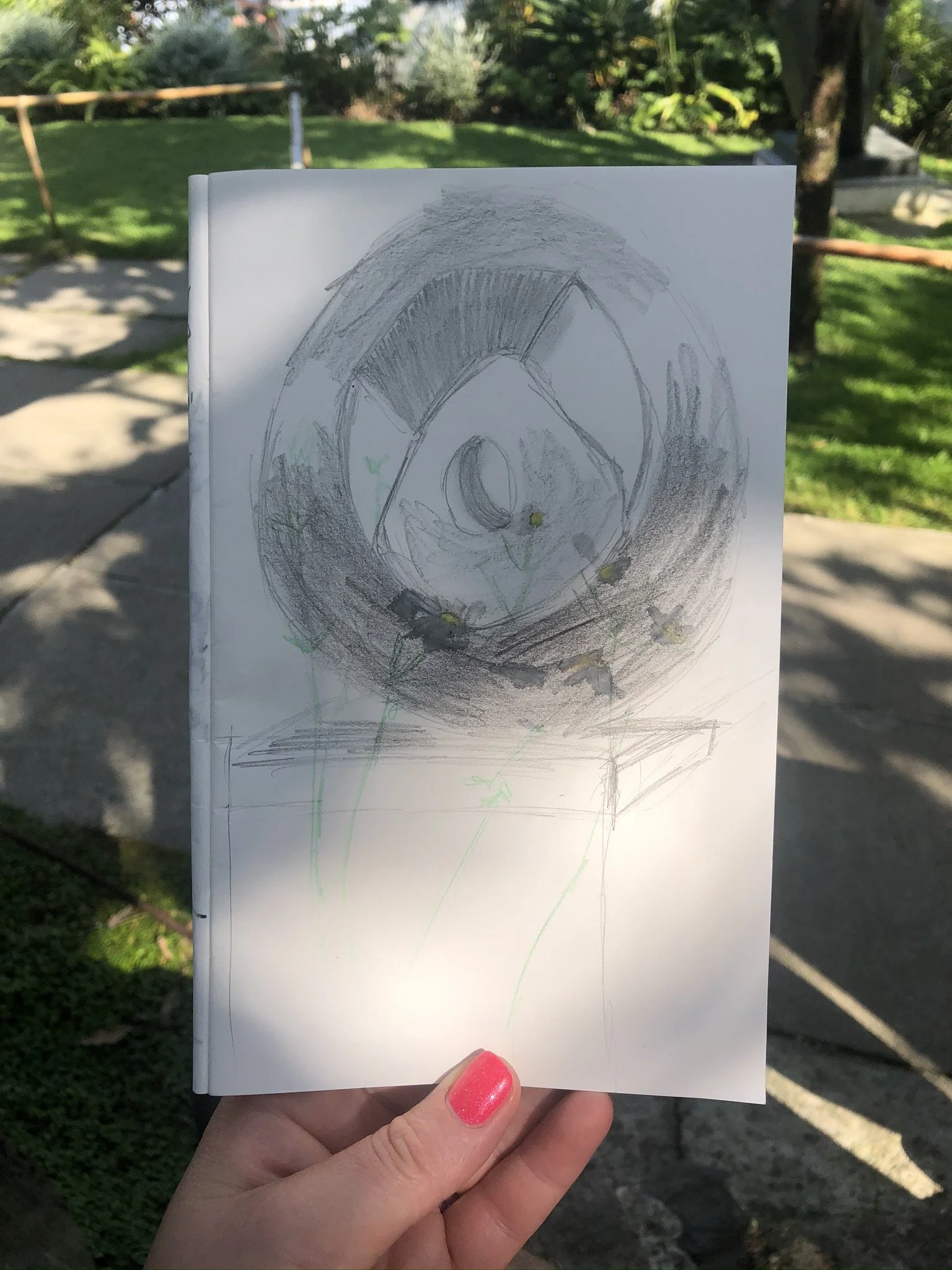 Sketch from Barbara Hepworths garden. A large round sculpture with a hag stone like form in the middle. with daisies in the from. Sketchbook is partly in the shade. Held by a hand with a pink polished thumb nail.
