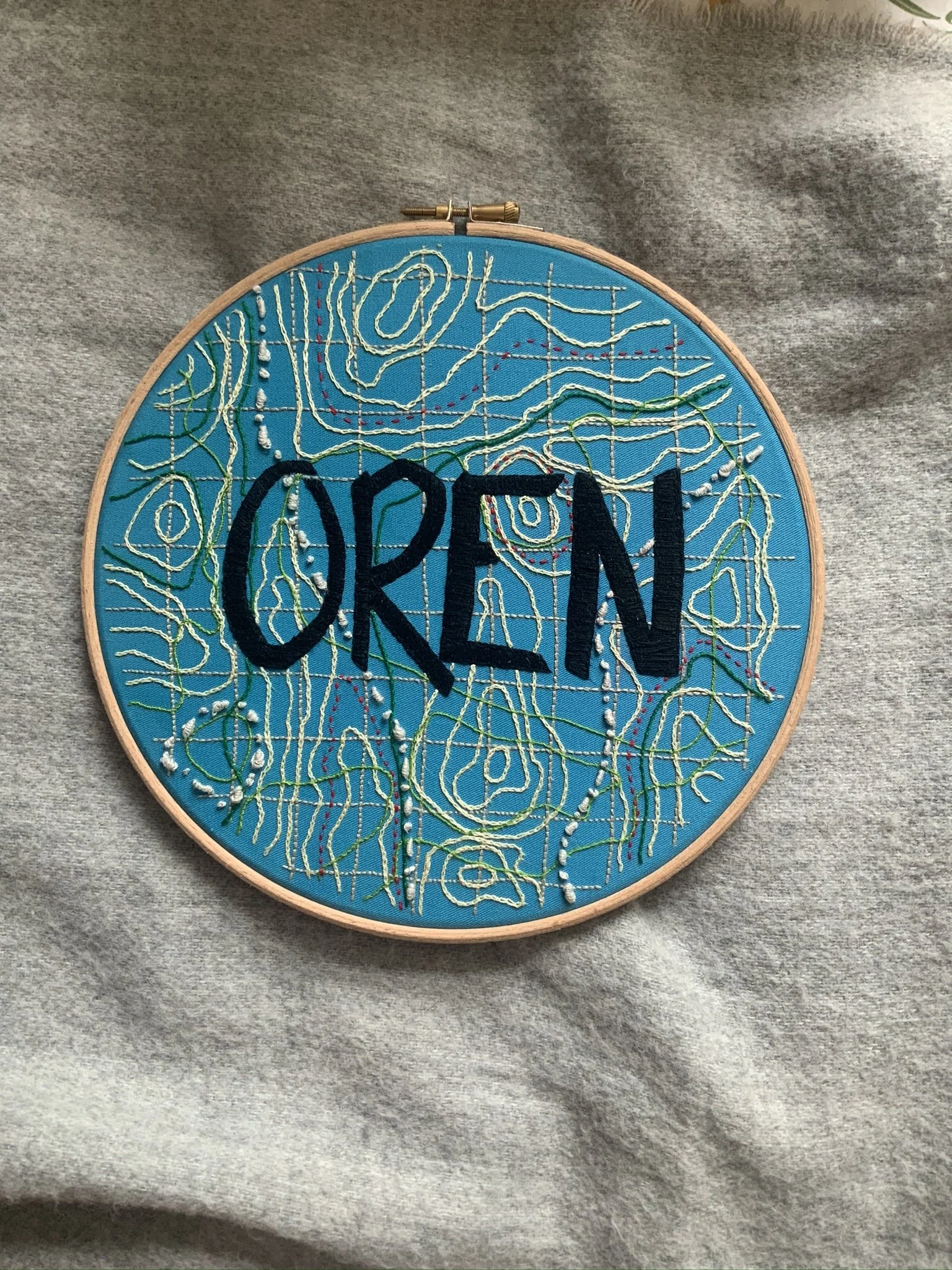 Embroidery hoop with a blue background, featuring the word 'OPEN' stitched in black, with abstract topographic map outlines in various colors and patterns.