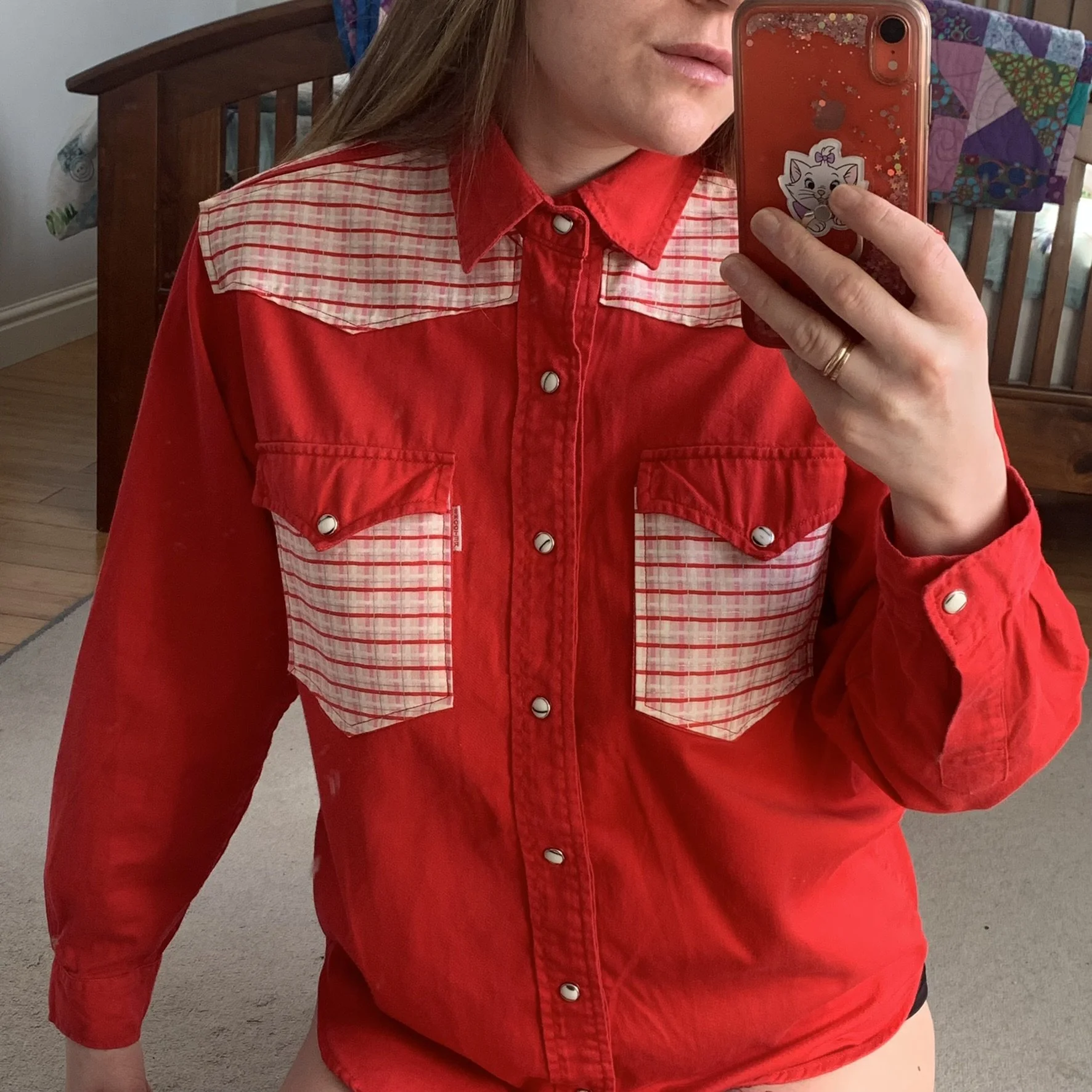 A person taking a mirror selfie wearing a red and white plaid western-style shirt with snap buttons and flap pockets. The collar area and pockets have been appliqued with delicate red white and pink gingham. Background of a bed and quilt.