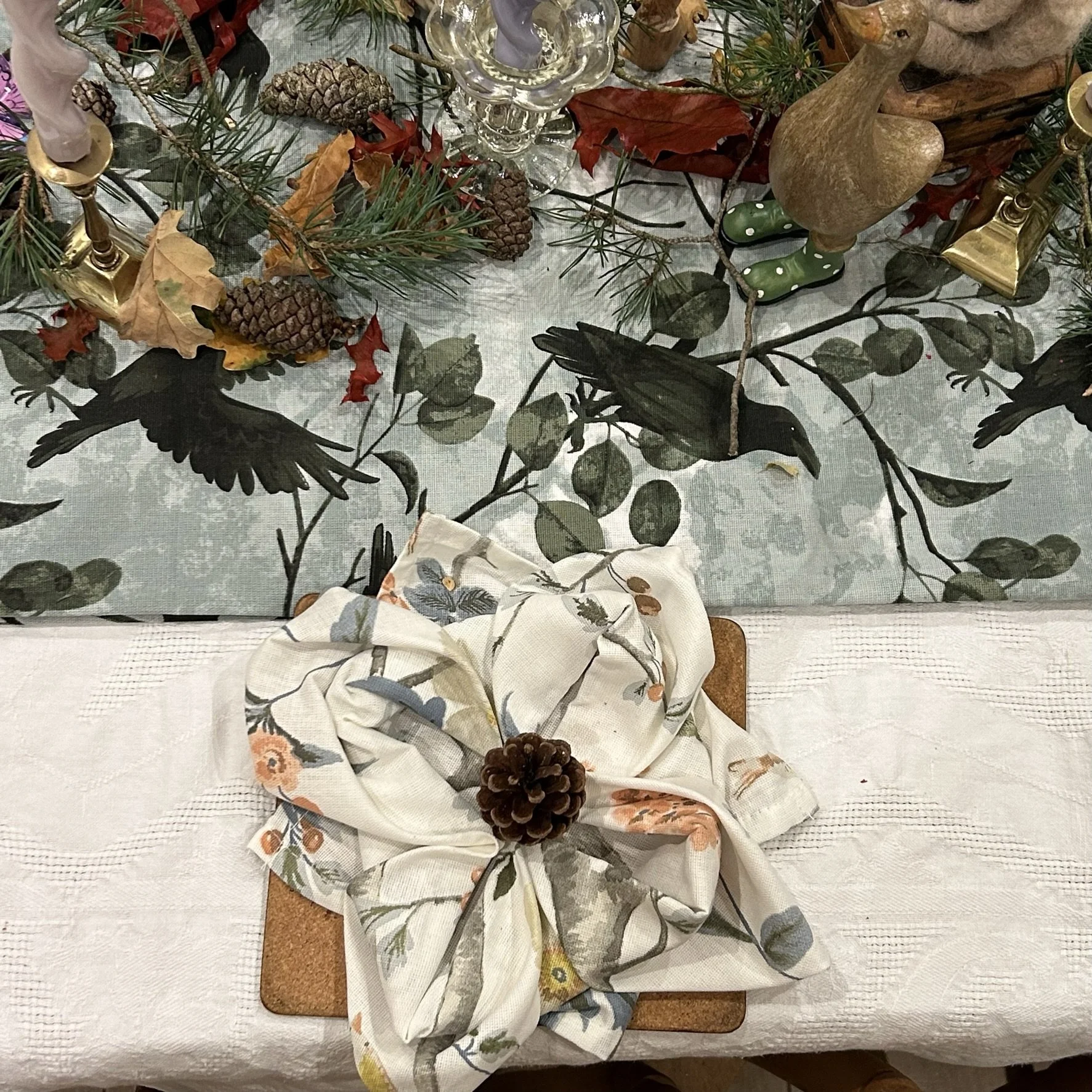 Table decorated for autumn with a tablecloth featuring black bird silhouettes and leafy branches, pinecones, dried leaves, greenery, and candles. A fabric napkin with a floral pattern is folded into a flat 4 petal flower with a decorative pinecone. 