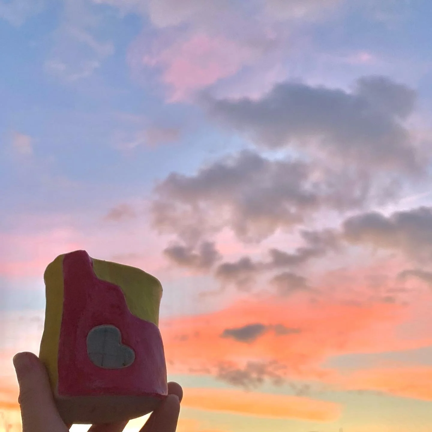 A hand holding a colorful pot with pink, yellow, and green surface against a sunset sky with clouds.