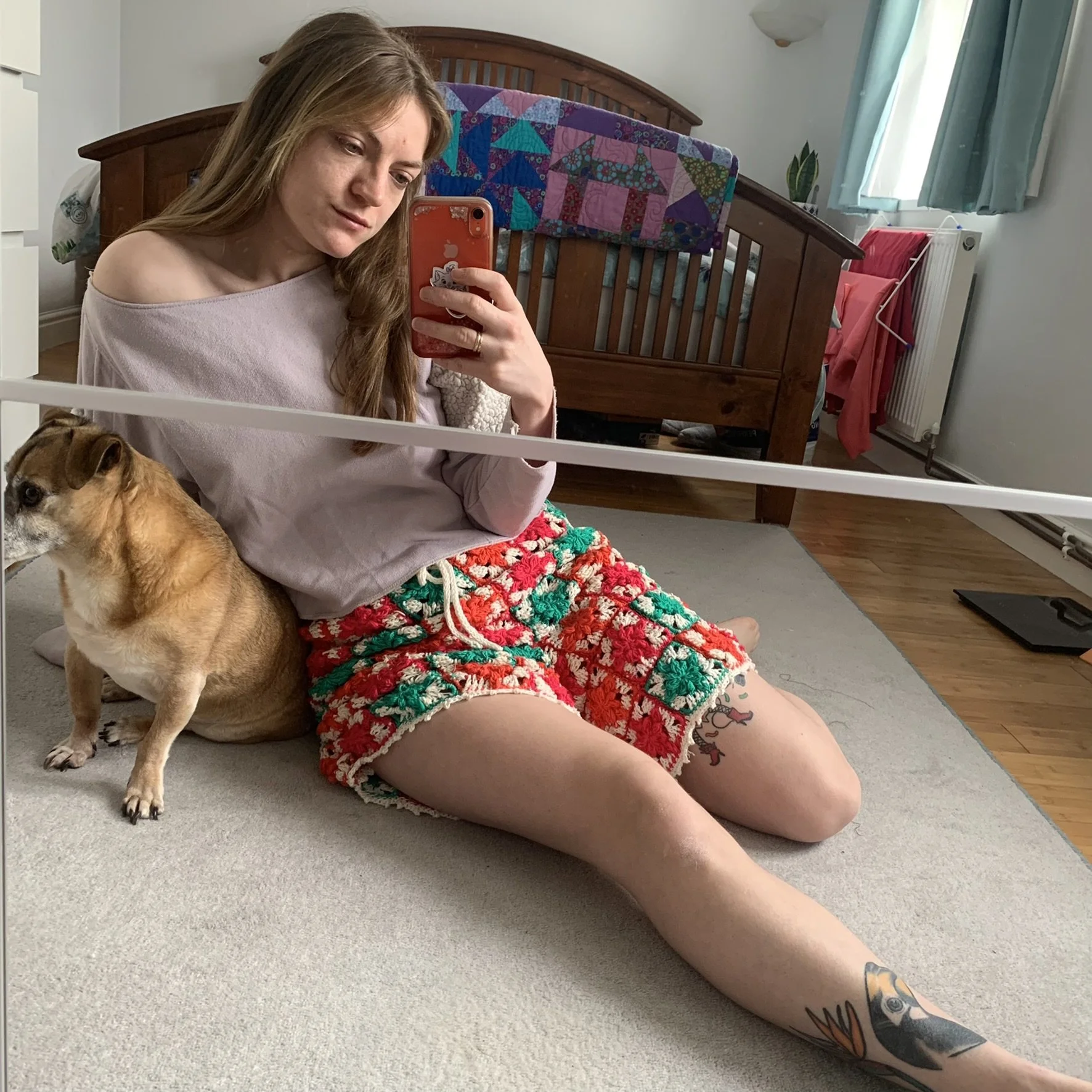 long hair, wearing a beige top and colorful crocheted shorts, taking a selfie in  mirror on the floor. She is sitting with her legs extended, and a dog is leaning against her. The room has a wooden bed with a colorful quilt, a window with teal curta