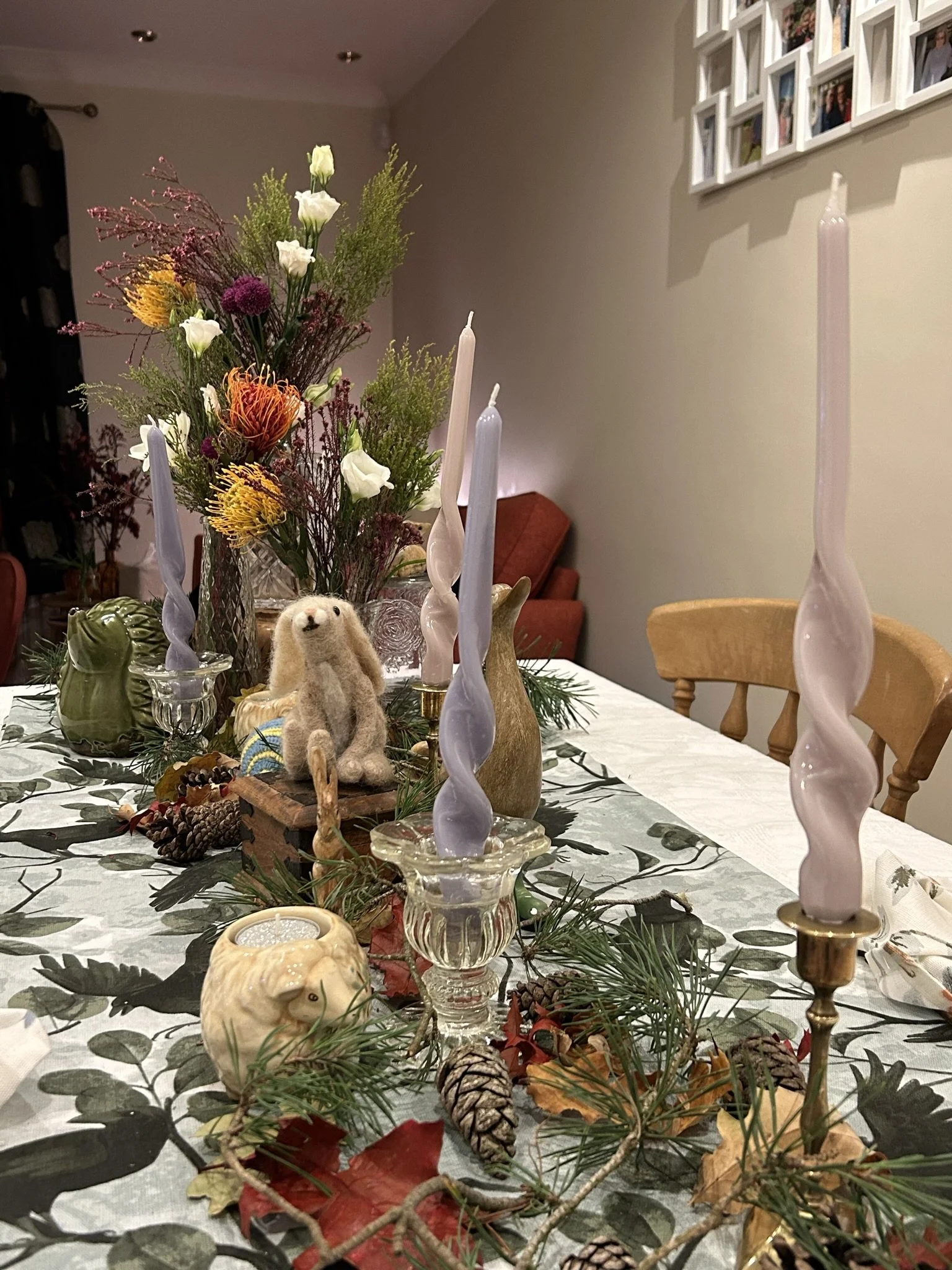 A decorated dining table with a floral centerpiece, pink and white twisted candles, pinecones, and autumn leaves, with a wall of framed photos in the background.