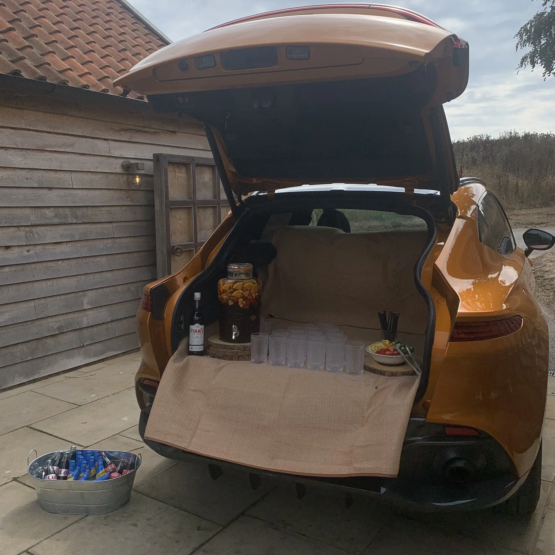 Open trunk of a yellow car with cups, a bottle, a glass jar of fruit, and a bowl with lemon slices inside, set up for a Jaguar brand event. A bucket of bottles is on the ground nearby, and the background shows a wooden fence and outdoor landscape.