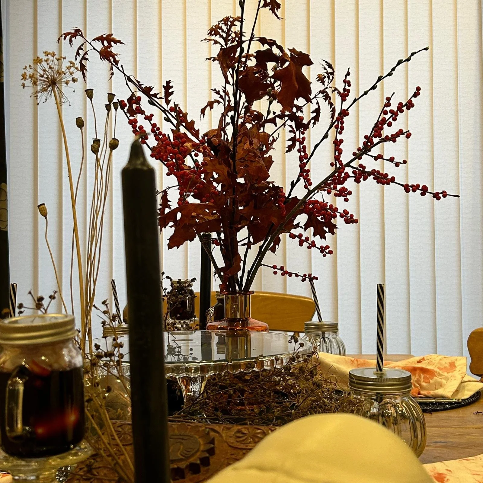 A Halloween centerpiece on a wooden dining table featuring a bouquet of autumn leaves and red berries in a  glass vase, surrounded by jars and decorative items, with a backdrop of vertical blinds.